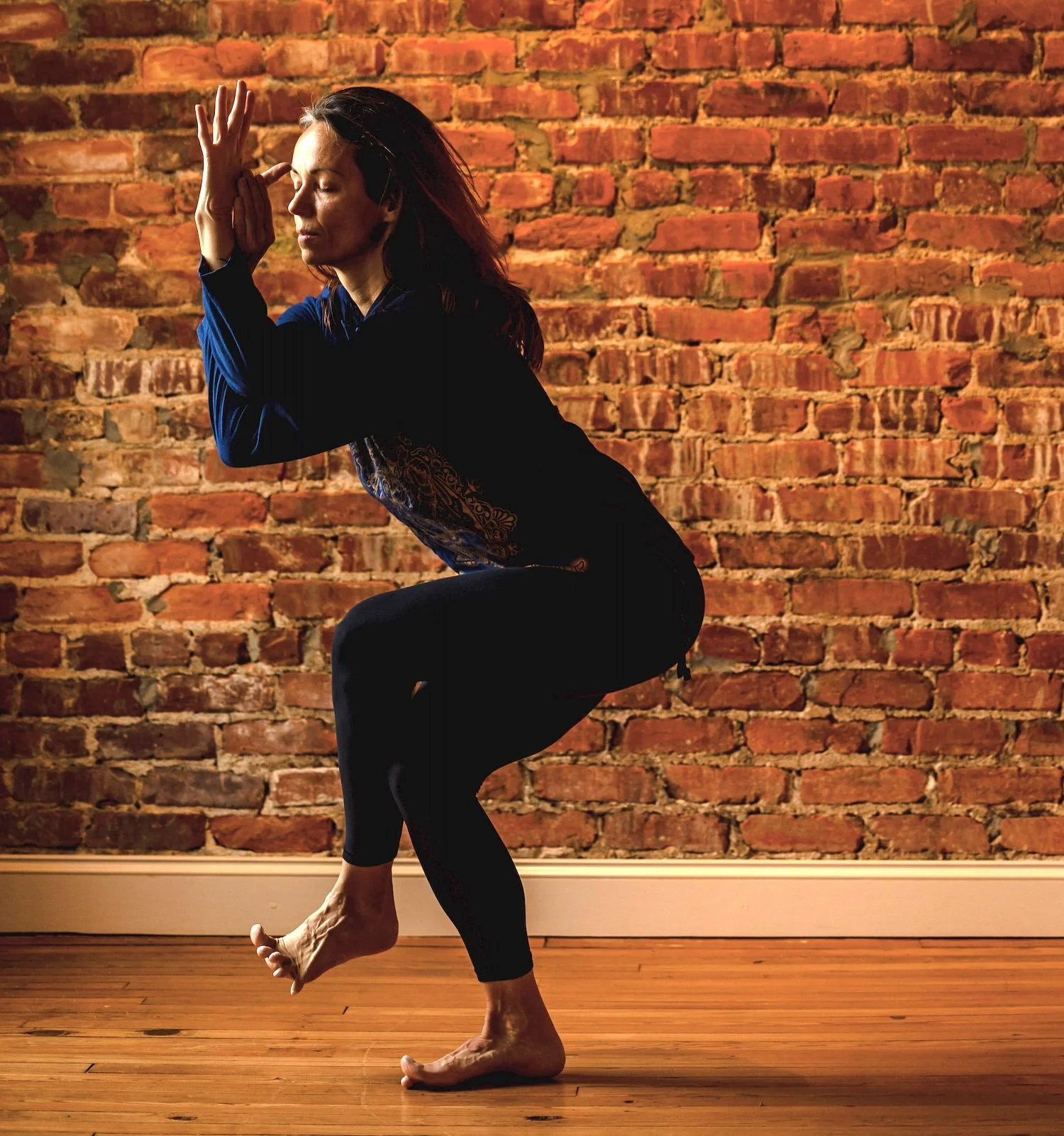 A woman practicing yoga indoors, balancing on one leg with the other leg bent and lifted, her hands in a prayer position near her face, in front of an exposed brick wall.