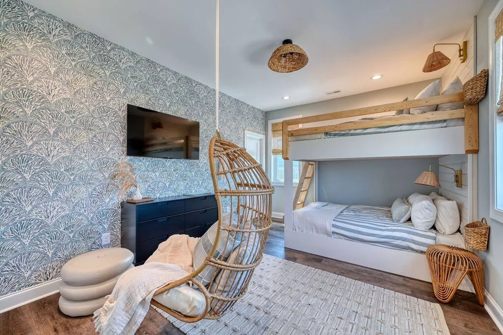 Beach-themed bedroom with a bunk bed, hanging egg chair, wall-mounted flat-screen TV, black dresser, and woven decor.