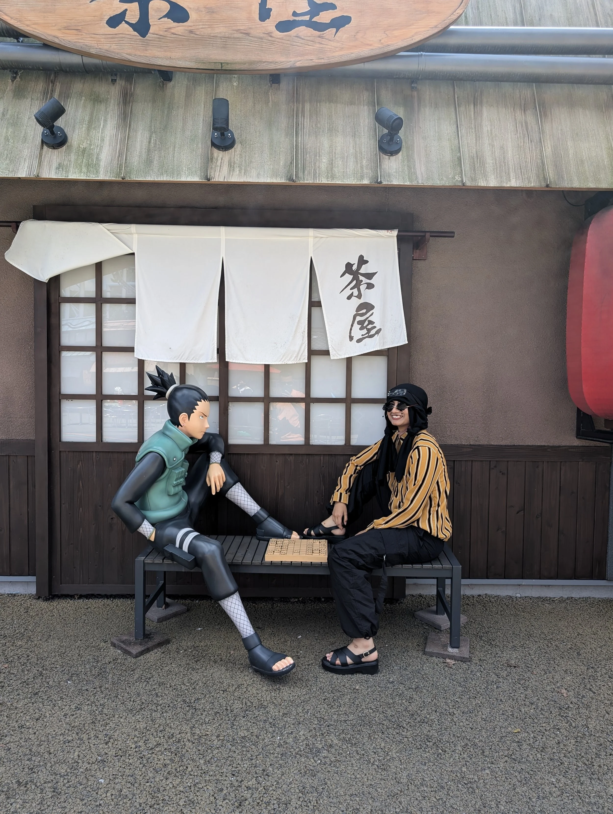 A woman wearing black sunglasses, a striped yellow and black shirt, and black pants, sitting on a bench, holding hands with a life-sized anime character statue of a male ninja from the anime Naruto, who is dressed in green and black, sitting on the bench. The background features a traditional Japanese-style window with white curtains, a white noren curtain with black Japanese characters, and a wooden sign with black characters. The ground is asphalt.