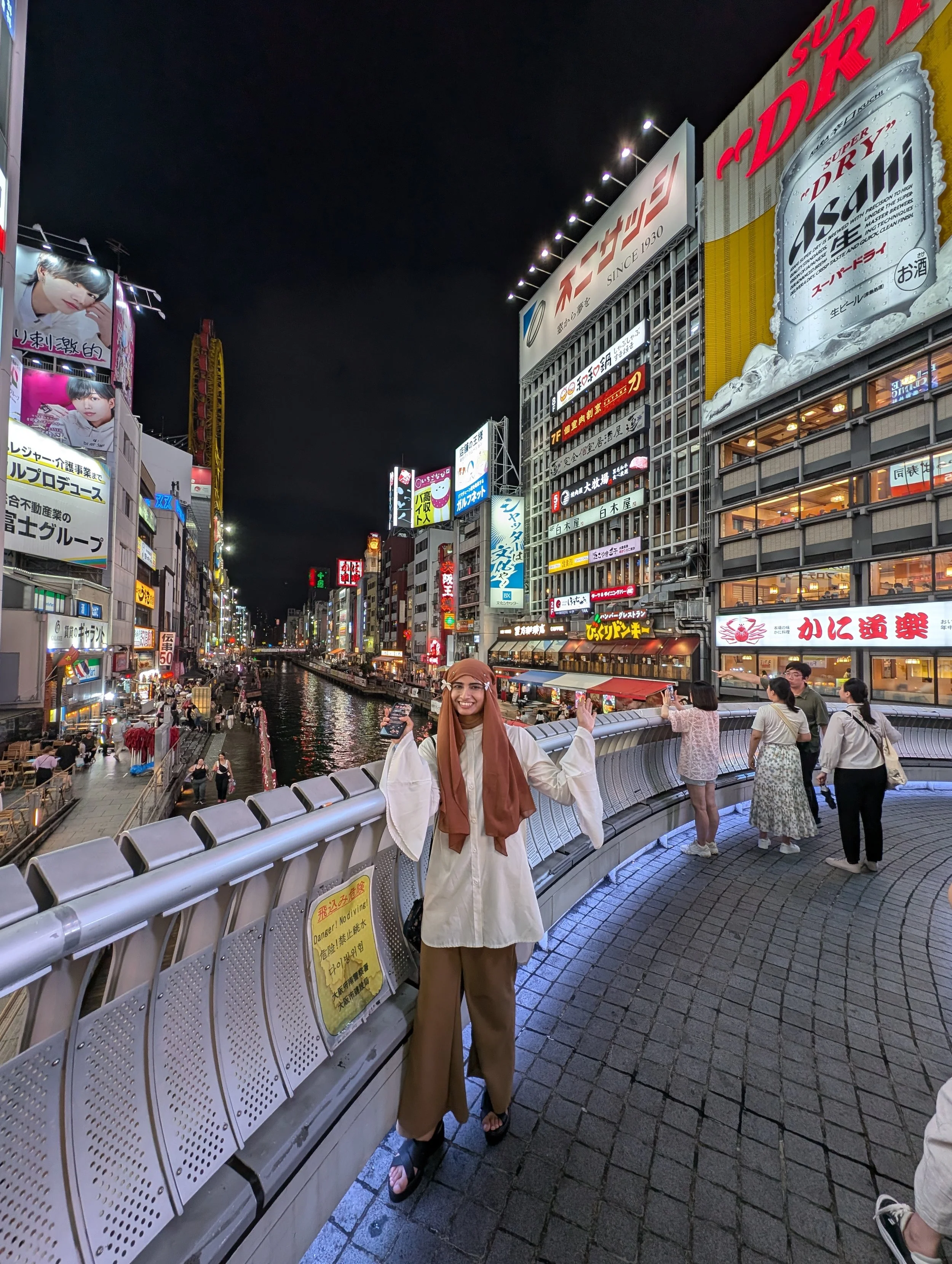 Night scene of a busy street with illuminated signs in Japanese, a canal, and people walking on a pedestrian bridge. A woman in the foreground is smiling and waving, wearing a beige jacket, brown pants, and a headscarf.