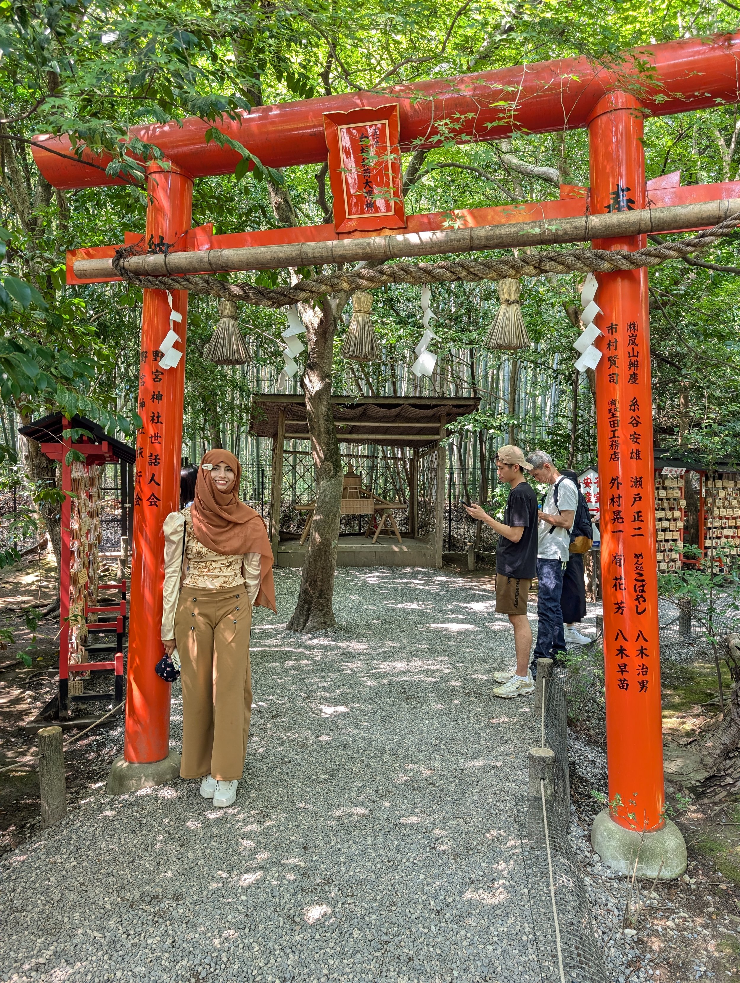 A red torii gate in a wooded area with three hanging shide paper streamers. Four people are standing underneath, including a woman smiling at the camera, and three others engaged with their phones.