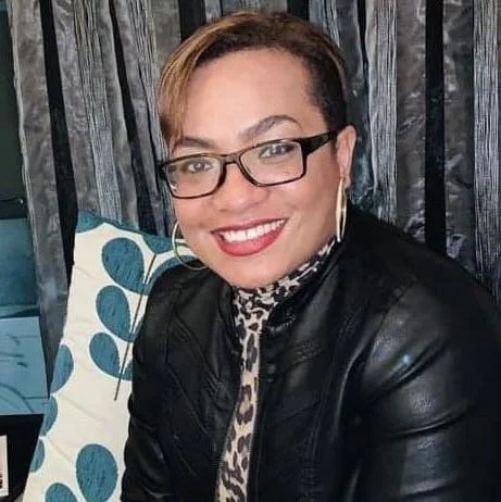 A brown Fijian woman with short hair, glasses, and a big smile, wearing a black leather jacket and leopard print top.