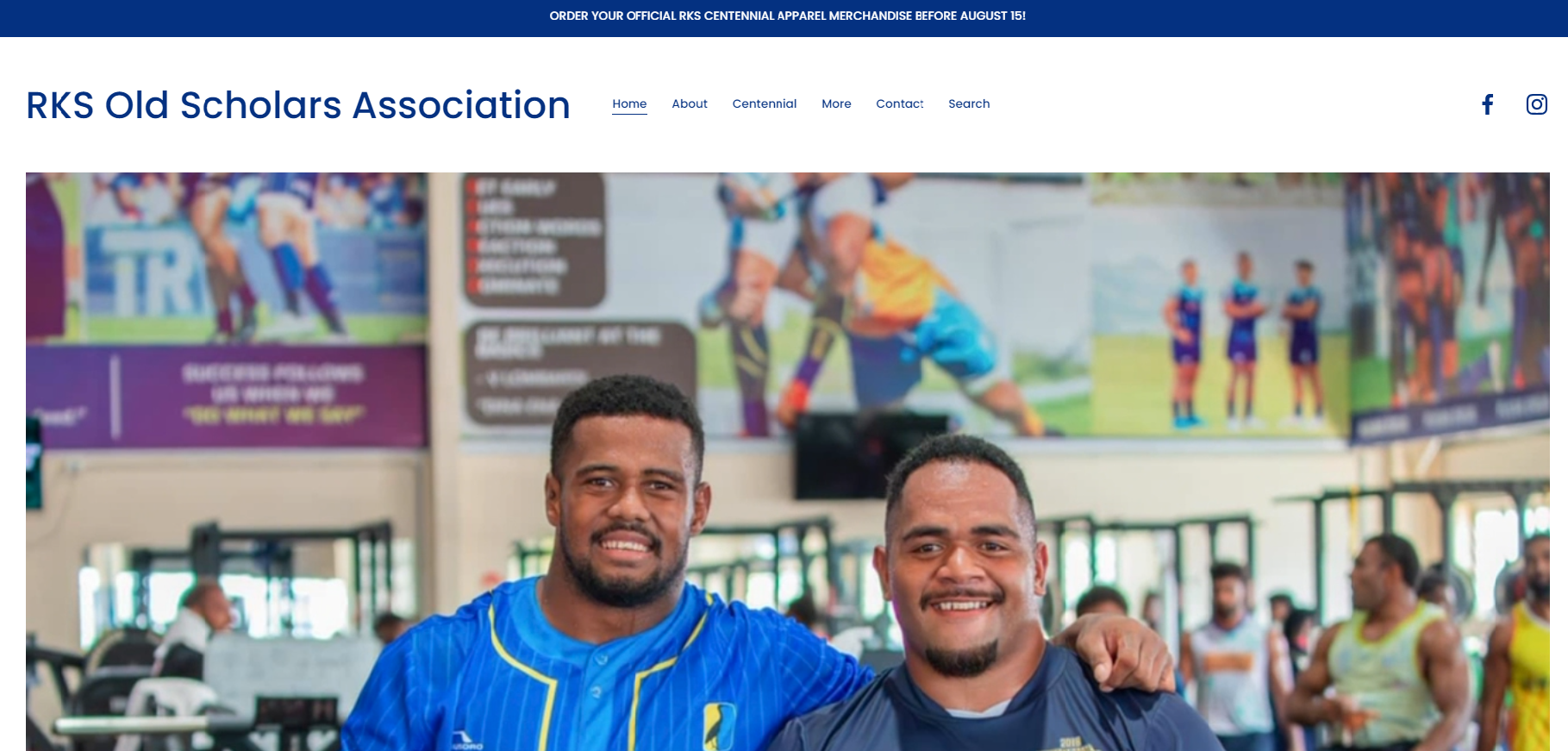 Two smiling men with athletic builds in sports attire, standing indoors at a sports facility or stadium. One man is wearing a blue jersey, and the other is wearing a dark sports shirt. The background shows other athletes, sports equipment, and large posters of rugby players on the wall.