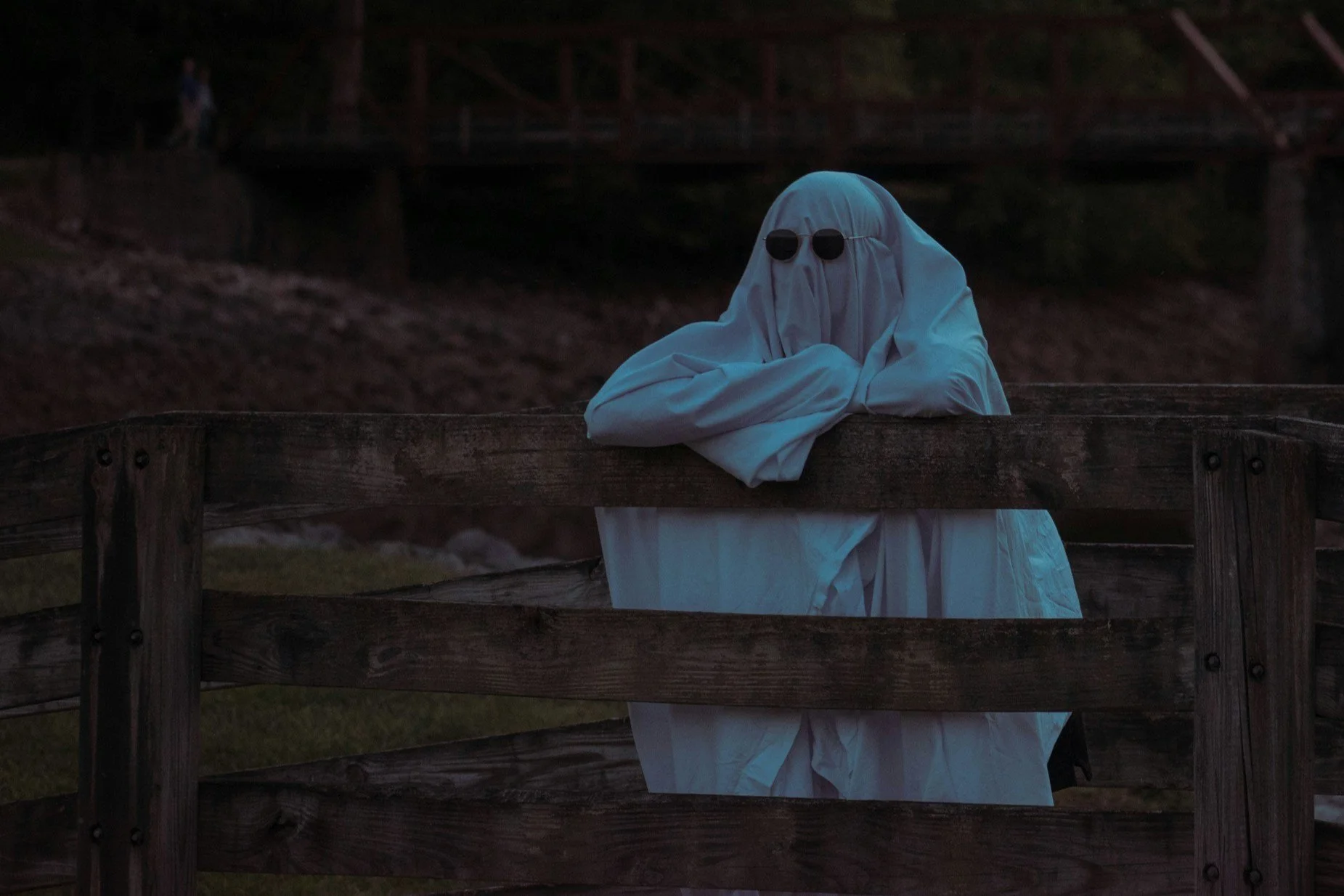 A person wearing a ghost costume with dark sunglasses leaning on a wooden fence during dusk or night.