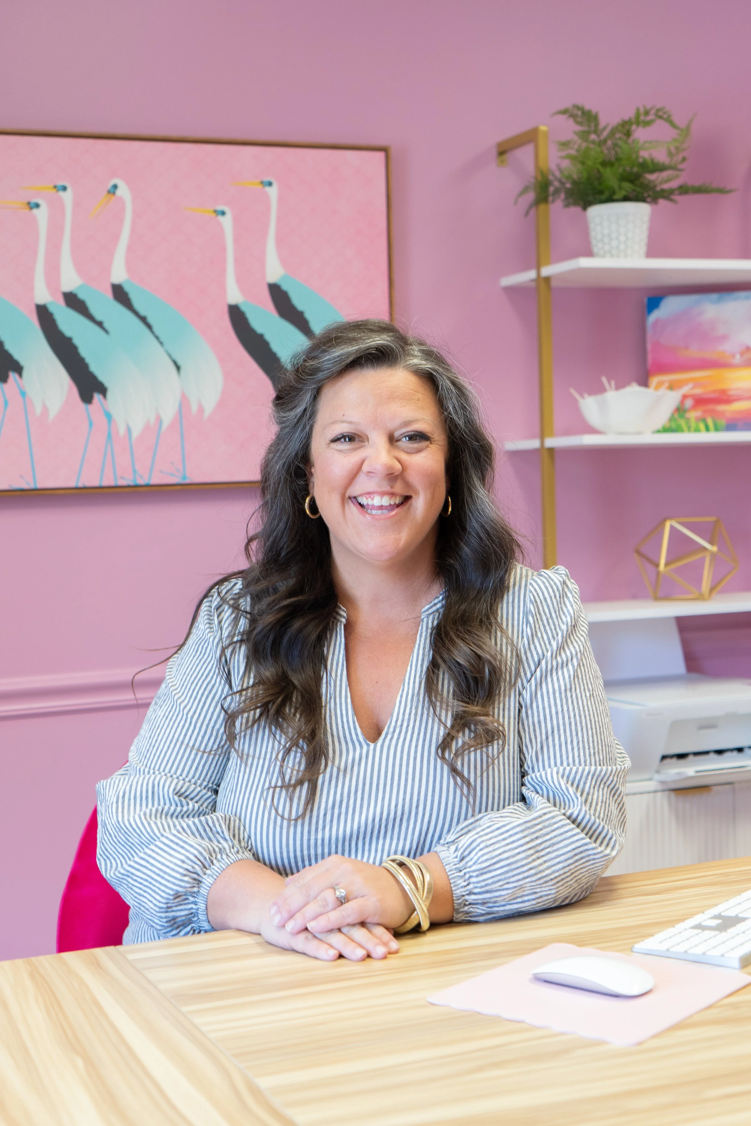 A smiling woman with long, curly dark hair, wearing a striped blouse, sitting at a wooden desk in a colorful office with pink walls, a painting of cranes, and decorative shelves with plants and artwork.