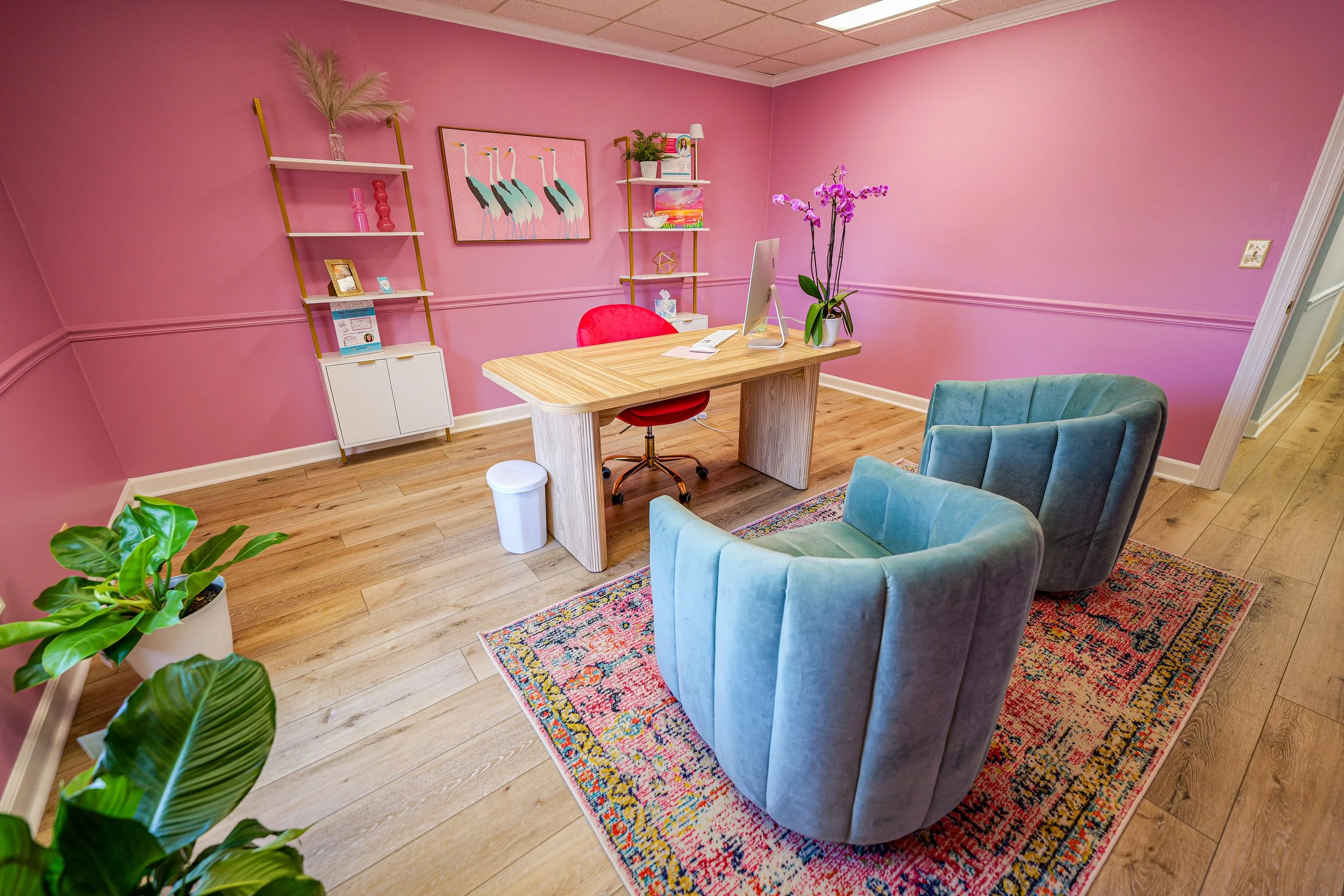 A colorful, cozy office with pink walls, a light wooden desk, two blue velvet armchairs, a patterned area rug, green plants, wall art of flamingos, and decorative shelves.