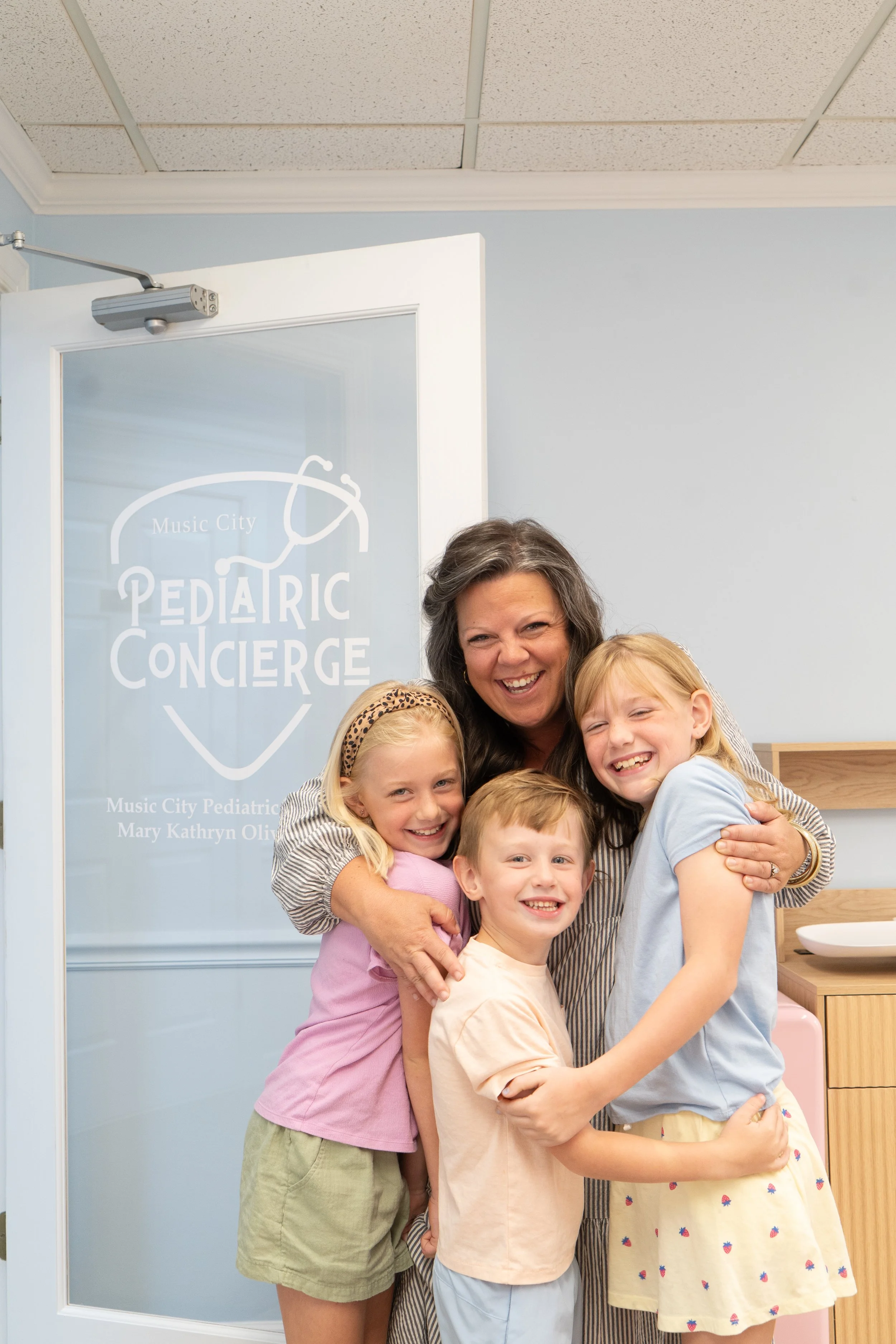 A woman and four children hugging and smiling in front of a door with a sign that reads 'Music City Pediatric Concierge' at a clinic.
