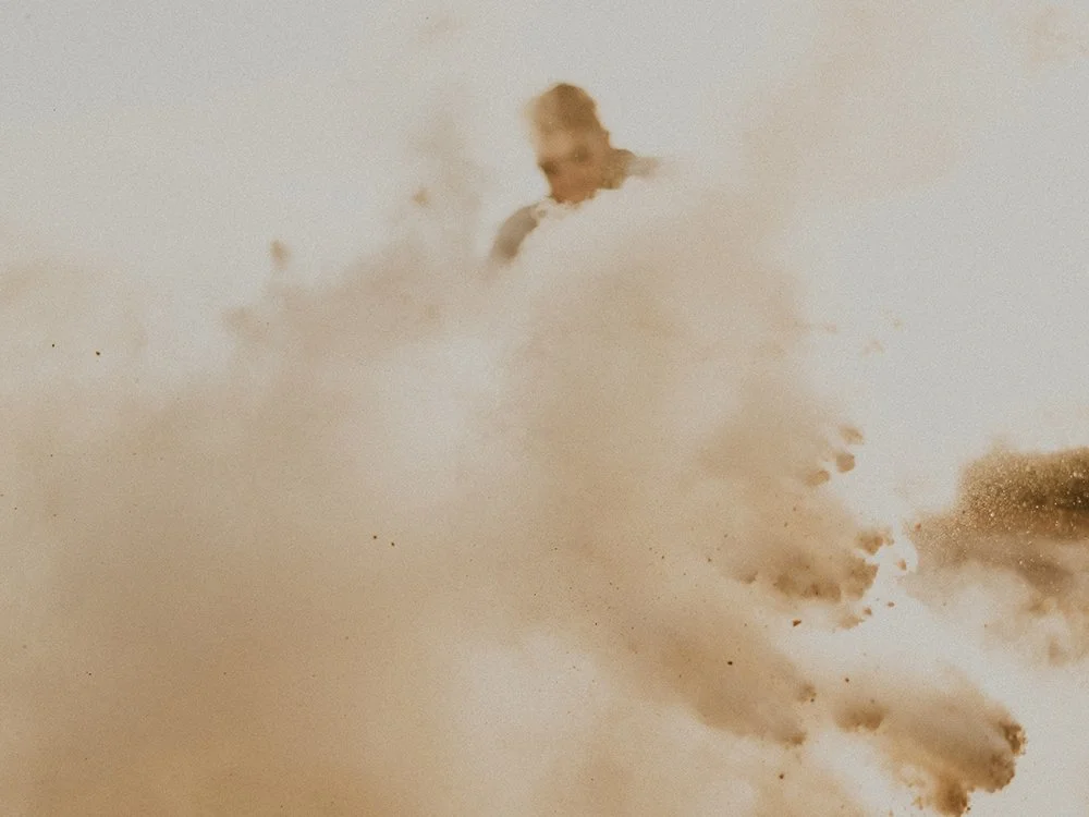 A person, partially obscured, emerges from a large cloud of dust or smoke in a bright, beige-colored environment.