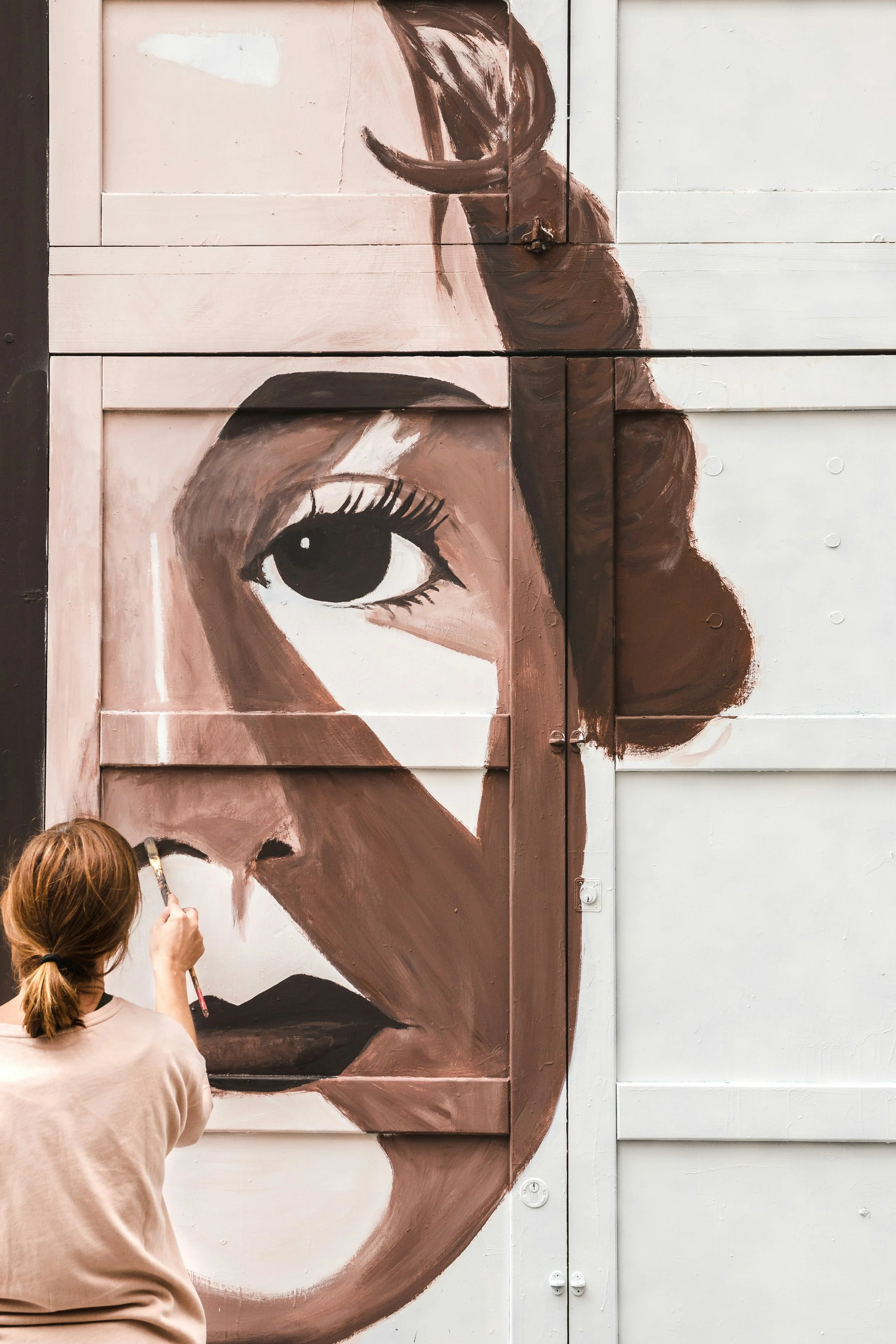 A woman painting a large portrait of a woman's face with detailed eye, nose, and lips on a wooden wall.