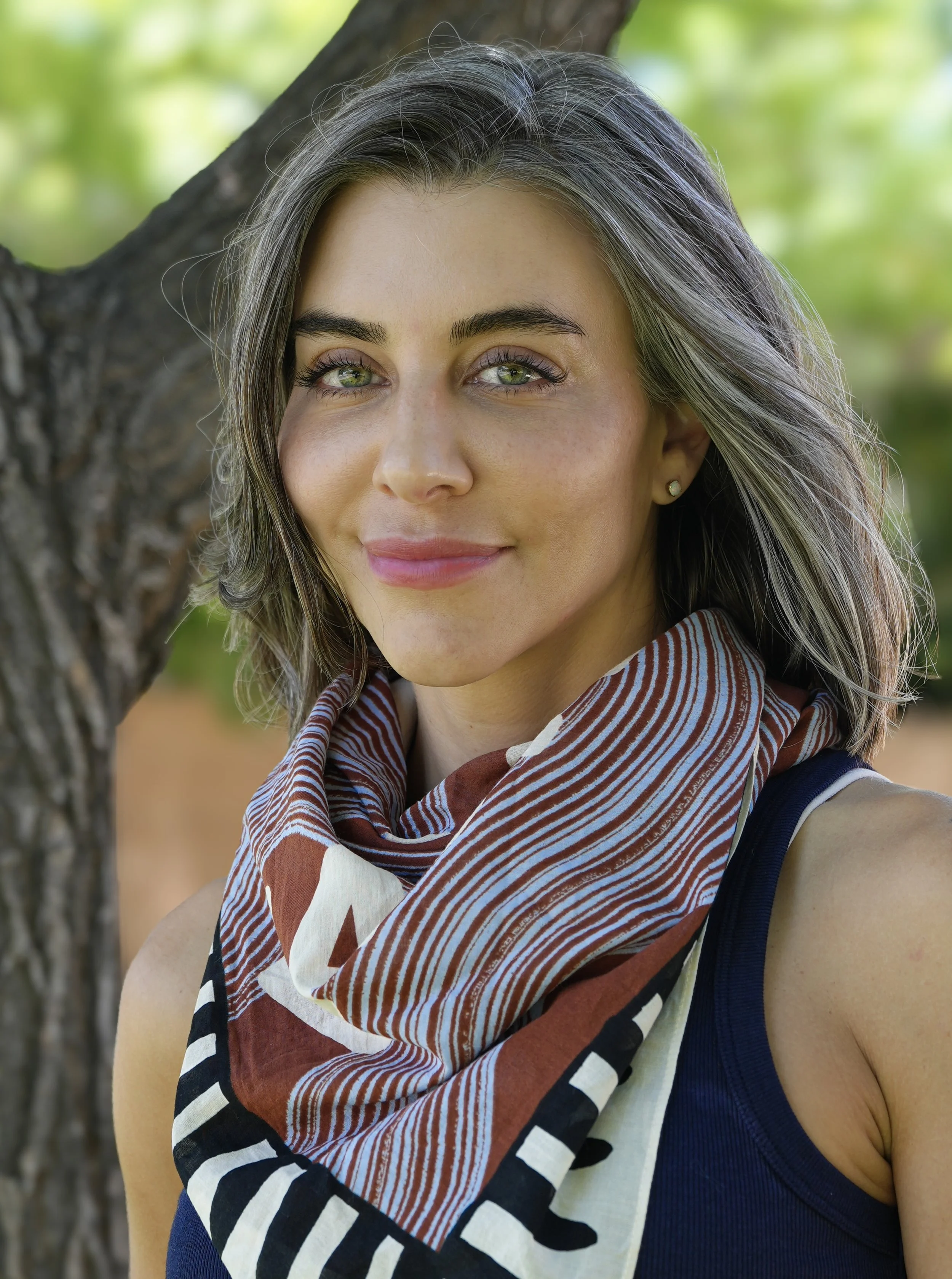 Close-up of a woman with short, gray-highlighted hair leaning against a tree outdoors, wearing a striped scarf and sleeveless top, smiling softly.