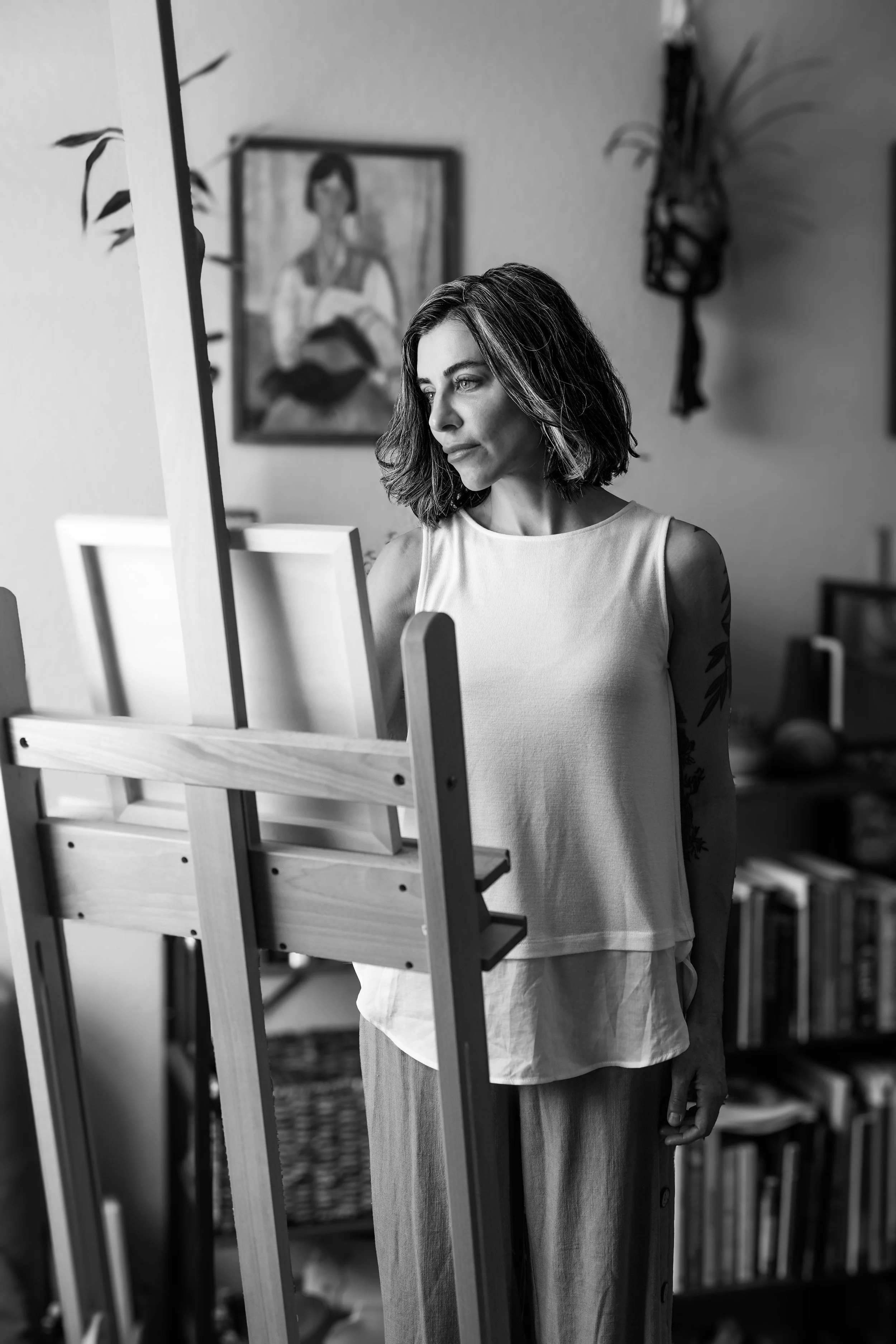 A woman with shoulder-length hair standing in front of an easel, looking at a canvas, in a room with artworks and books visible in the background.