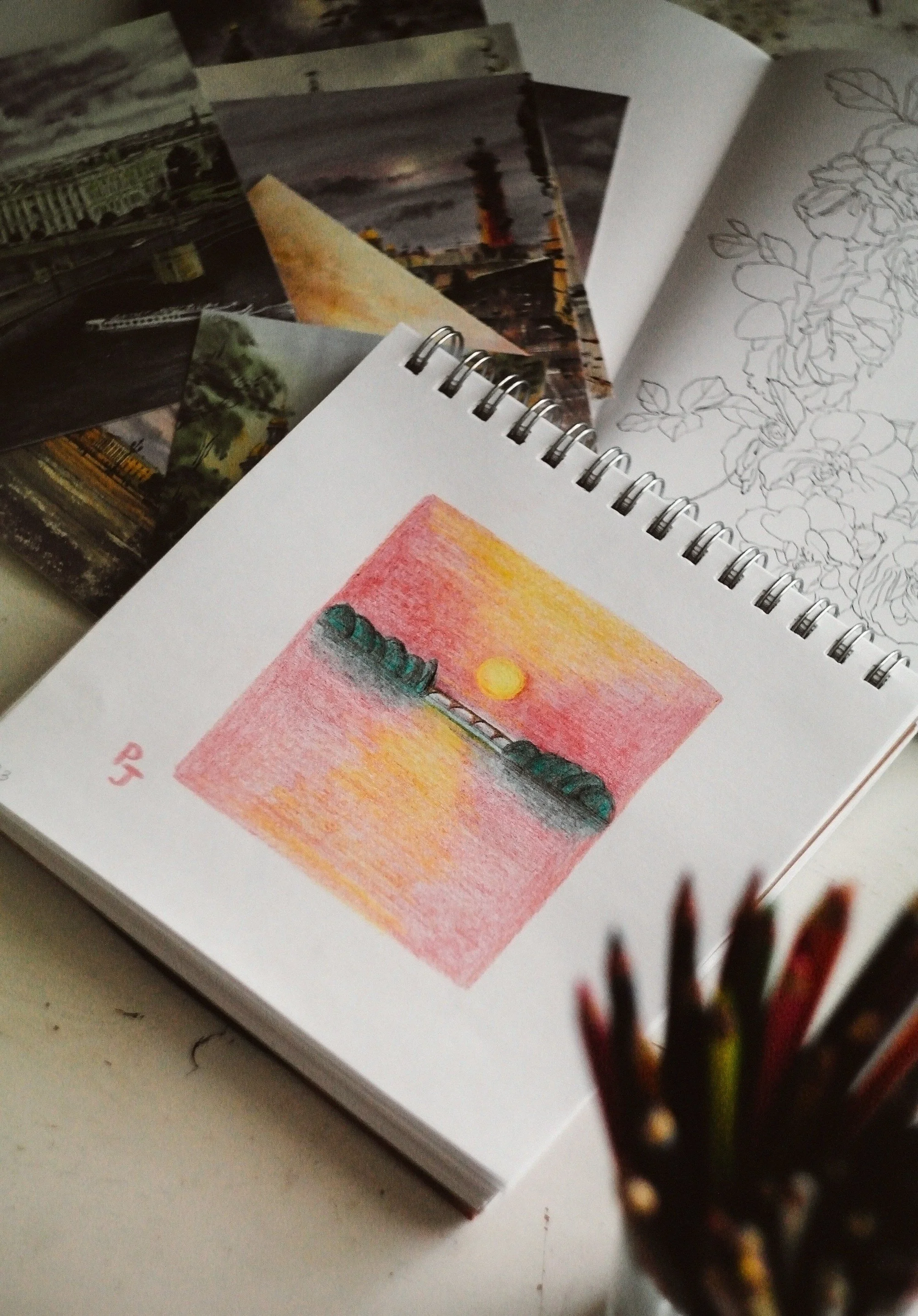 A spiral-bound sketchbook open to a colored pencil drawing of a sunset over water with a boat, and a black ink outline drawing of flowers on the adjacent page, with a blurred background of colored pencils.