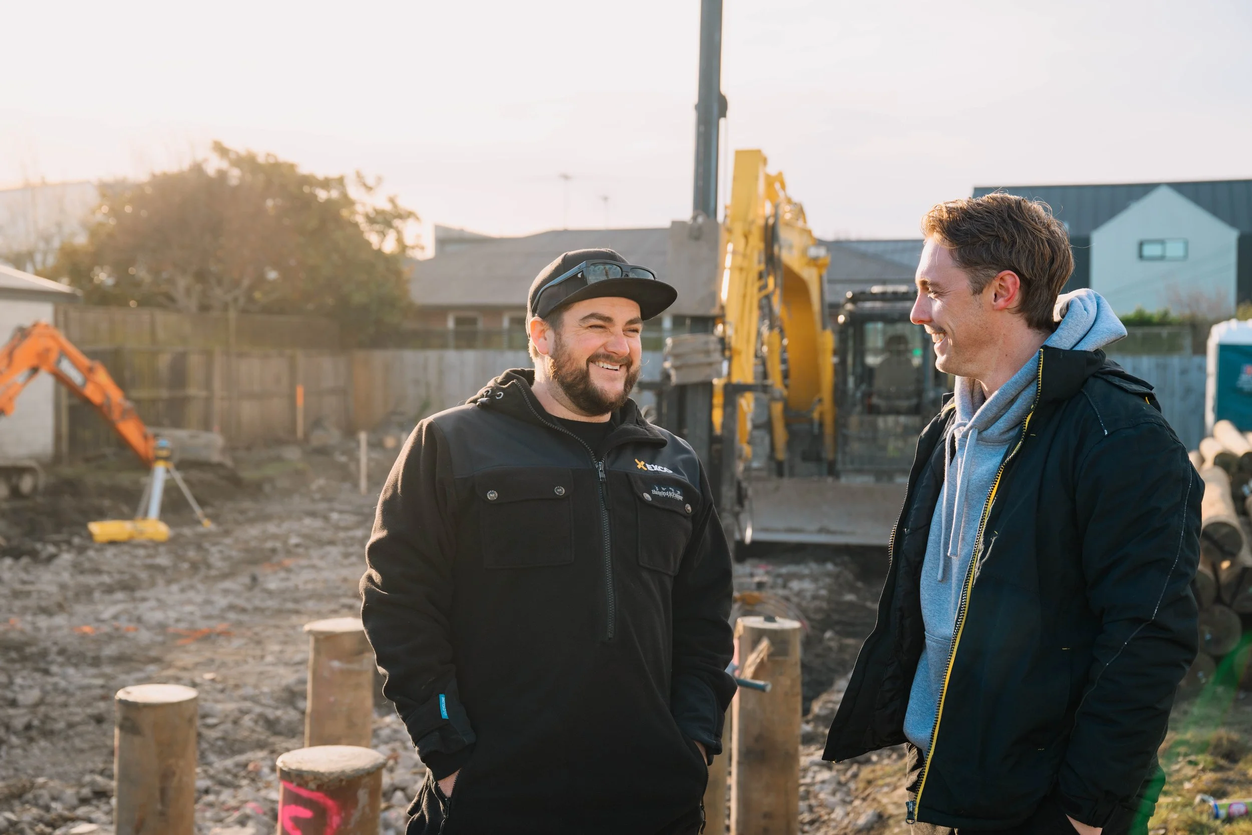 Two men are smiling and talking at a construction site with equipment and a partially built structure in the background, during daytime.