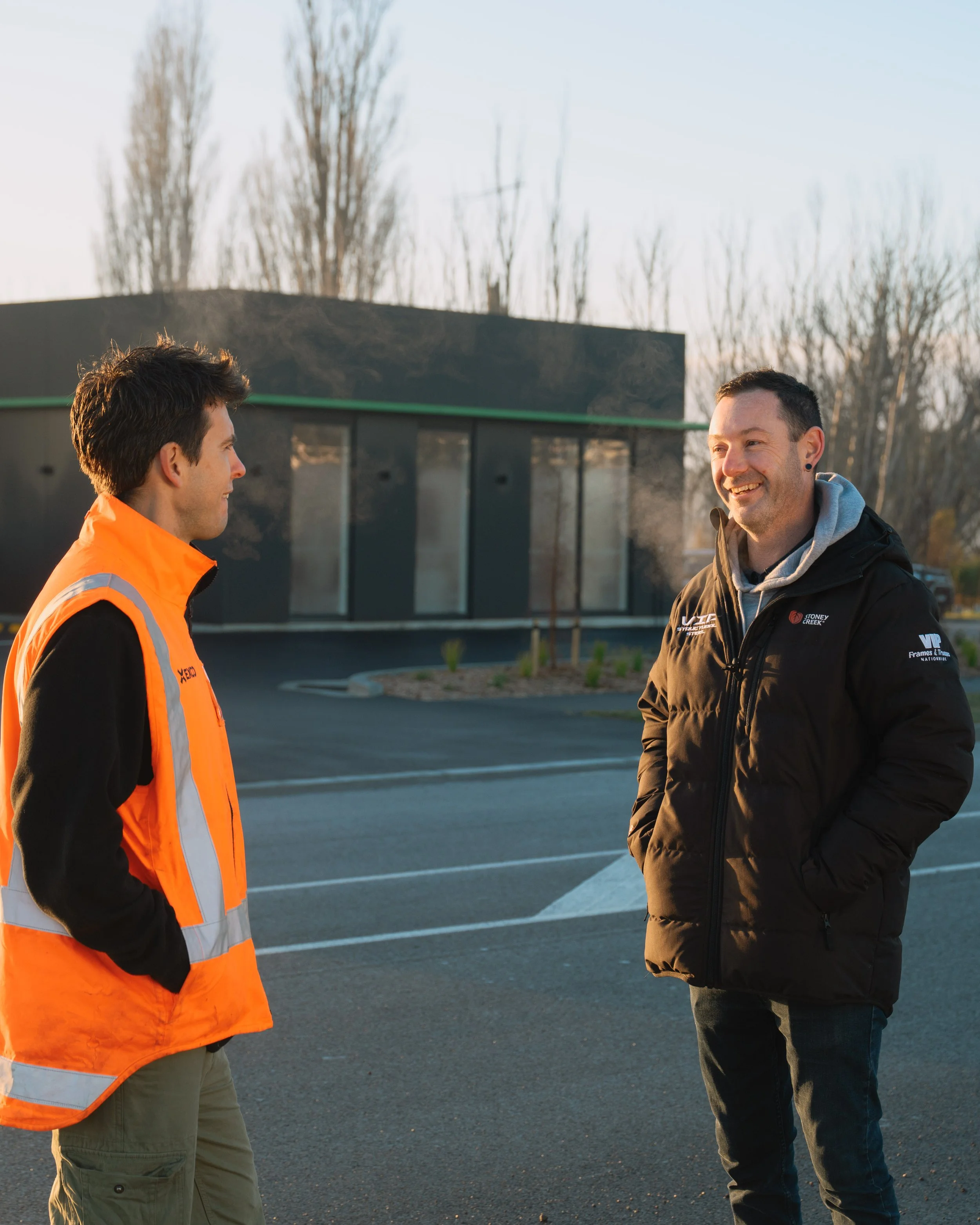 Two men are standing outdoors in a parking lot, engaged in conversation. One is wearing an orange safety vest and khaki pants, and the other is wearing a black jacket and jeans. The background features a building with large windows and leafless trees, indicating a late fall or winter setting.