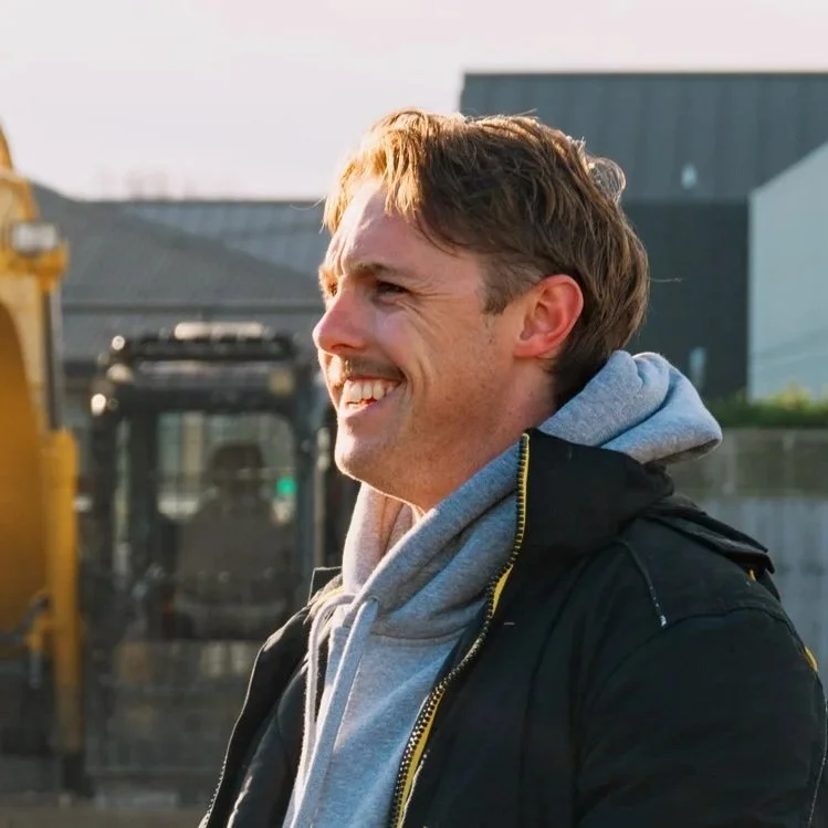 A man smiling outdoors, wearing a gray hoodie and black jacket.