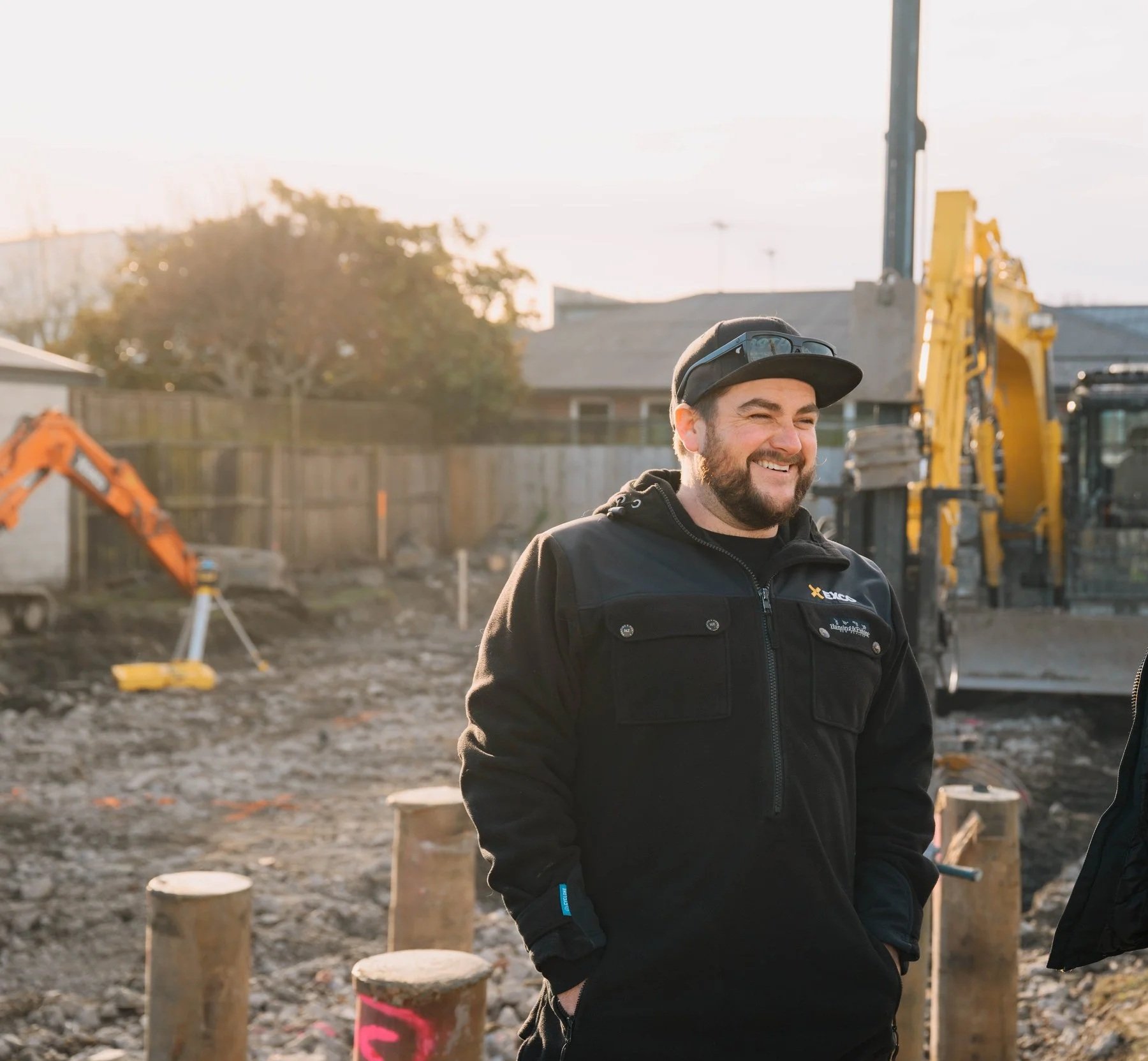 A man with a beard and glasses on his head, wearing a black jacket, smiling outdoors on a construction site with machinery and wooden posts in the background during sunset.