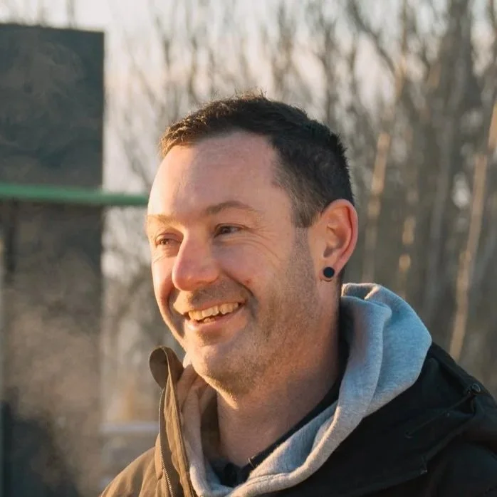 A smiling man with short dark hair, a beard, black earrings, wearing a gray hoodie and black jacket, outdoors with trees in the background.