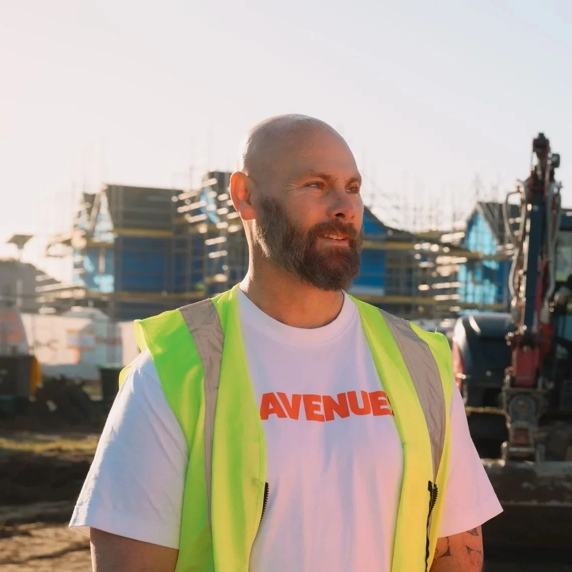 A bearded man wearing a white T-shirt with orange text and a yellow safety vest stands outdoors at a construction site with buildings and machinery in the background.