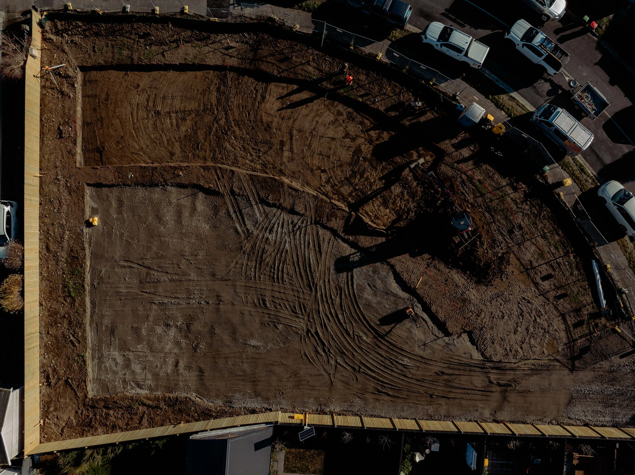 An aerial view of a construction site with a rectangular dirt area, surrounded by parked trucks and construction equipment, bordered by a yellow pedestrian walkway and a street with parked cars.
