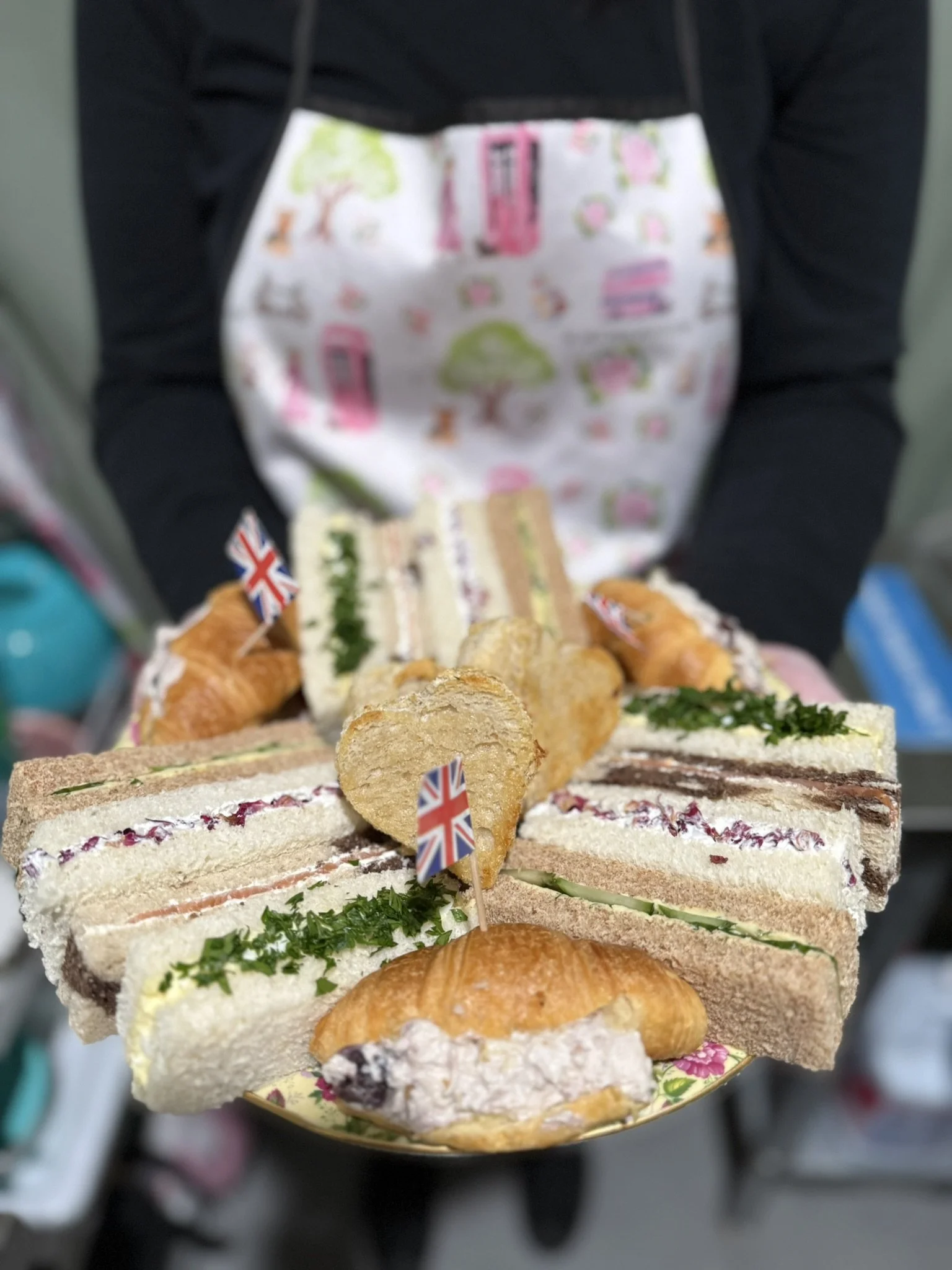 Tray of assorted sandwiches, croissants, and finger foods with small British flags, held by a person wearing a black shirt and a white apron with pink, green, and orange trees and houses.