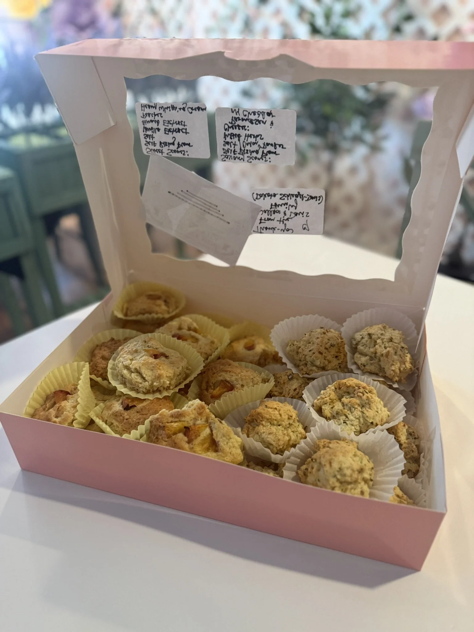 A pink box with a clear front, filled with assorted homemade cookies, some in white paper cups, on a white surface with a blurred outdoor background.