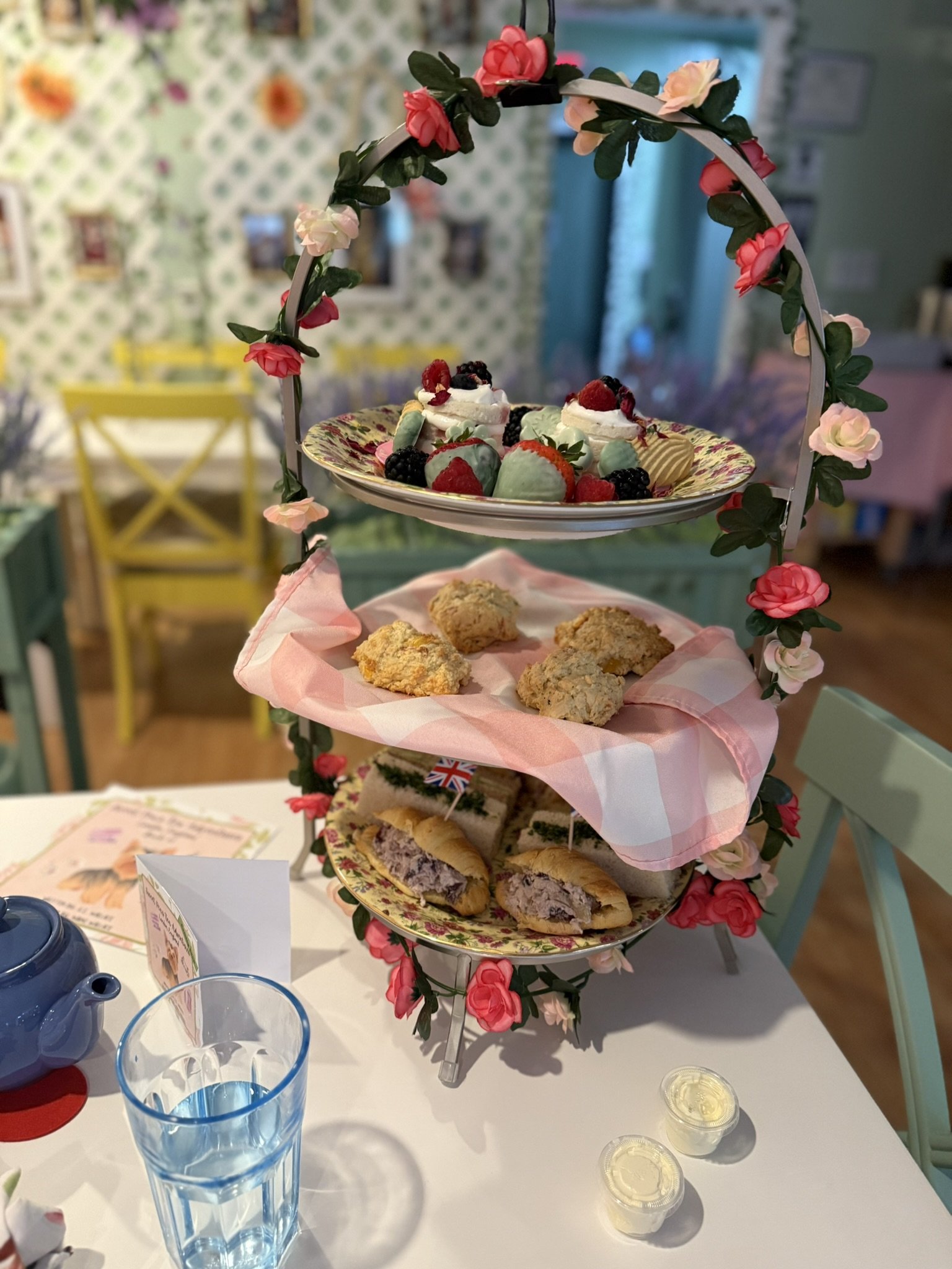 Three-tiered dessert stand decorated with pink and red flowers, holding assorted strawberries, berries, scones, and sandwiches, on a white table with a glass of water and teapot.
