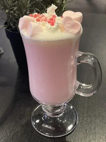 Pink hot chocolate topped with whipped cream and pink sprinkles in a clear glass mug