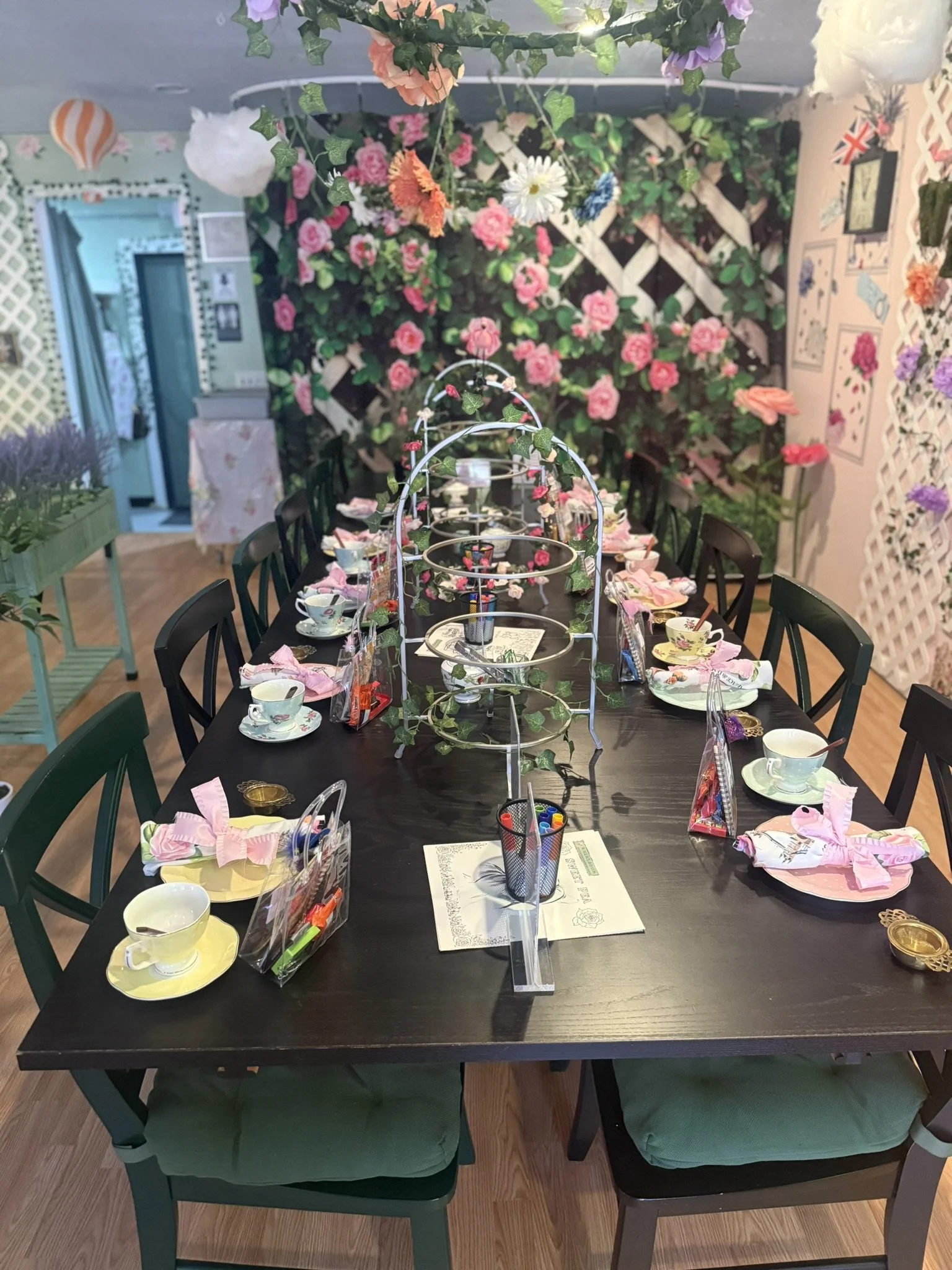 A decorated indoor table setup for a tea party with pink, purple, and white flowers, teacups, tea saucers, pink napkins with bows, and various party favors. The background features a wall covered with floral decorations and a lattice with flowers.