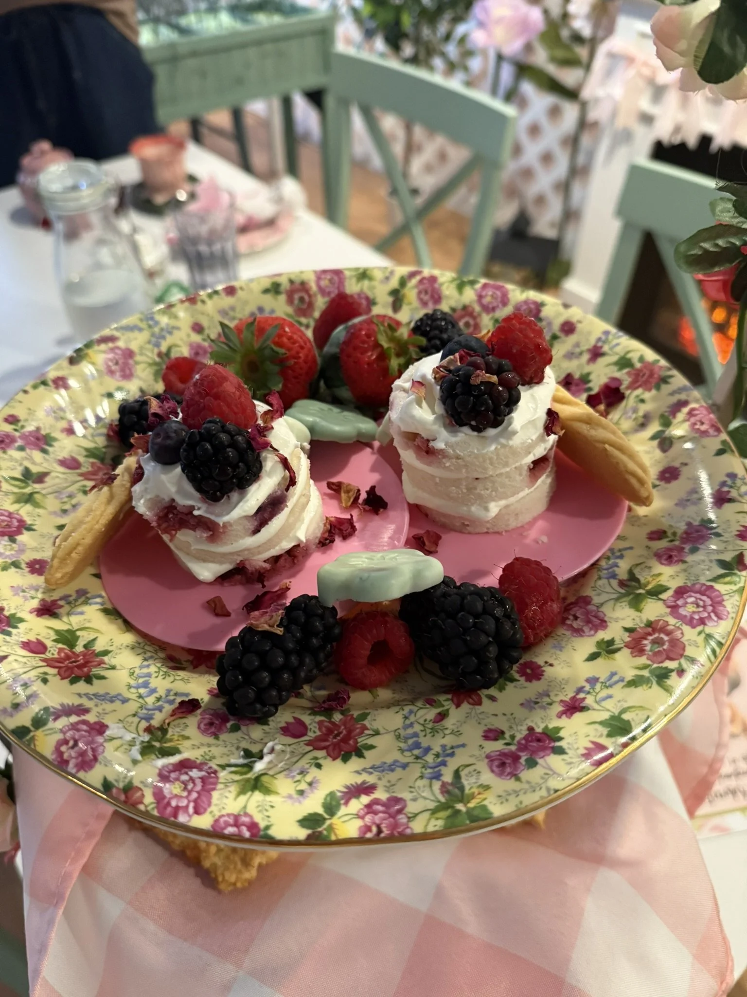 A decorative plate with two mini raspberry and blackberry desserts topped with whipped cream, surrounded by fresh berries, cookies, and mint leaves, on a table with floral and checkered tablecloths.