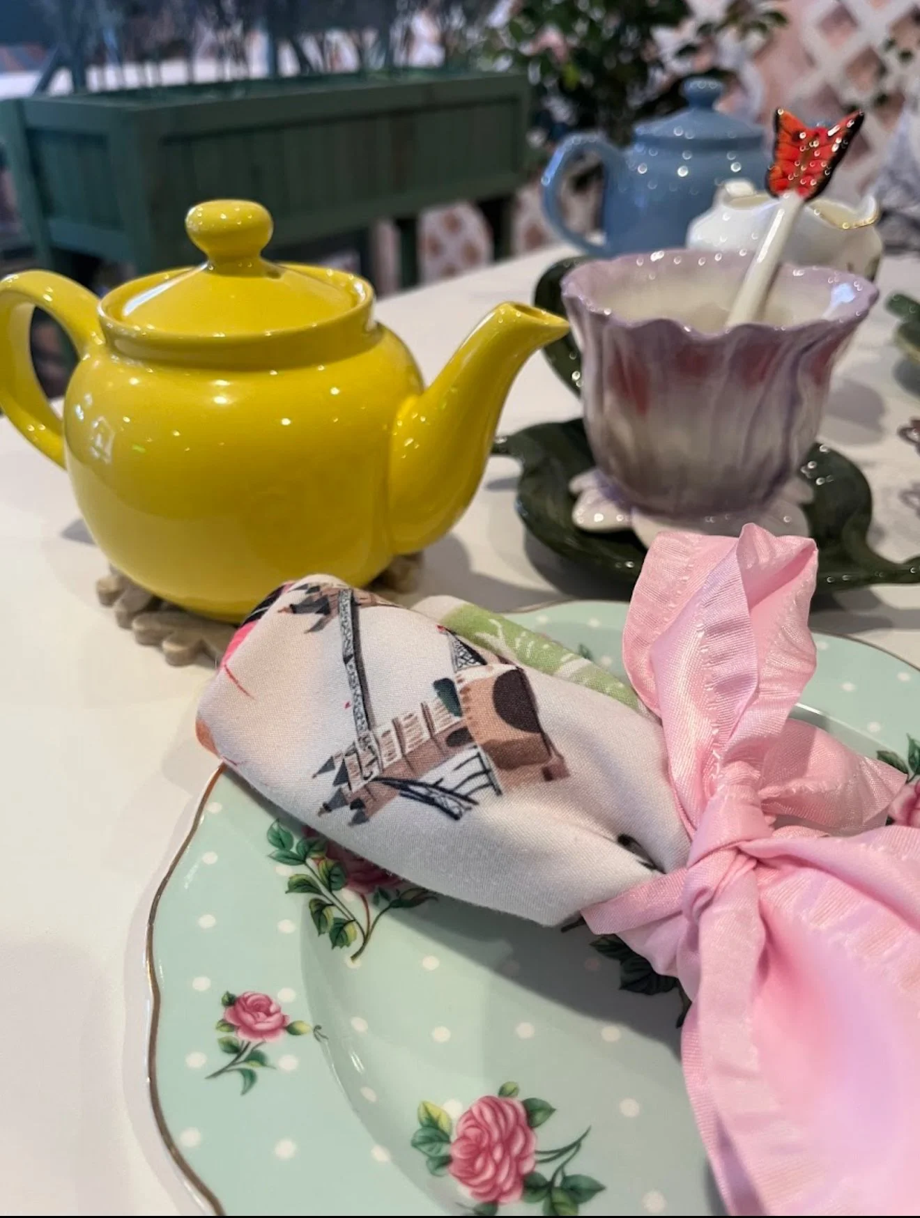 Colorful tea set on a table with a yellow teapot, a pink and purple marbled teacup with a butterfly-shaped stirrer, a green and pink floral plate with a wrapped cloth, and a tray with decorative elements.