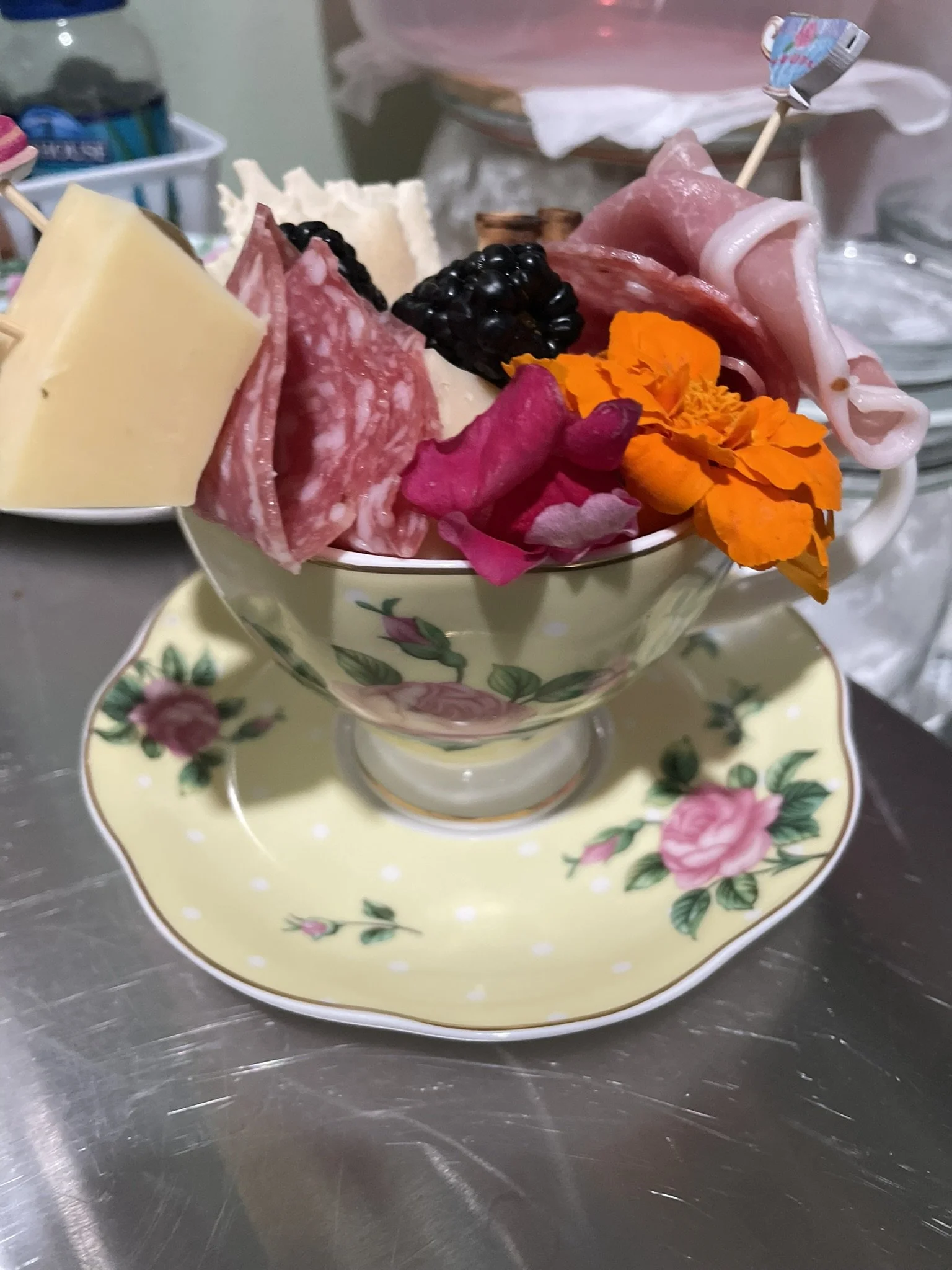 A decorative teacup filled with an assortment of cheeses, cured meats, blackberries, and edible flowers, placed on a matching floral saucer.