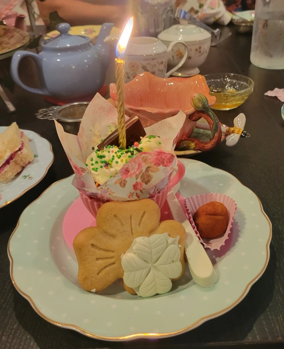 A birthday dessert plate with cookies, a cupcake with a lit birthday candle, and decorative items on a fancy plate; background shows teapots, cups, and other tableware.