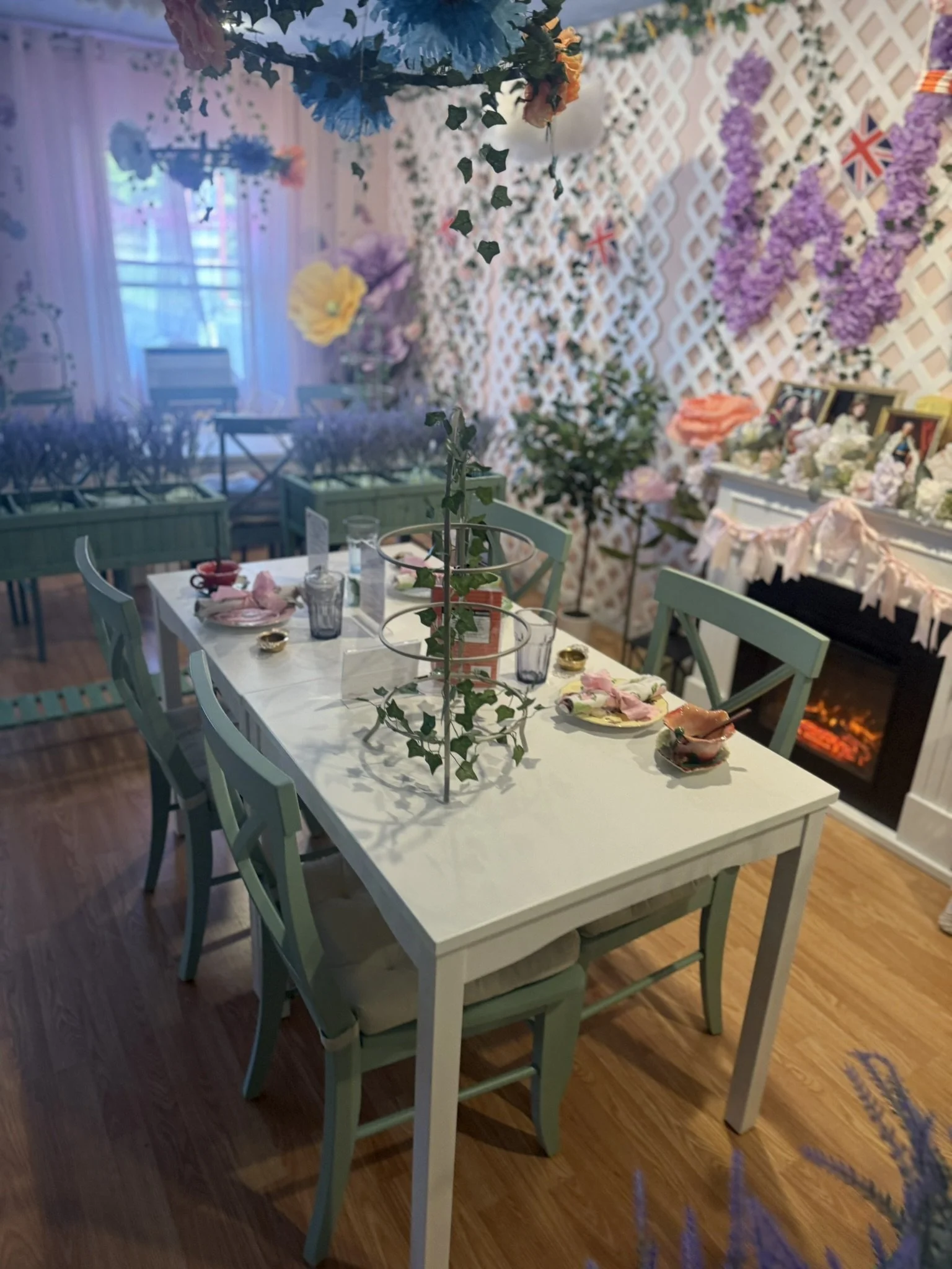 A decorated dining area with a white table and pastel green chairs, set with plates, glasses, and napkins, in a room decorated with flowers, paper hearts, and Union Jack flags, suggesting a festive or patriotic celebration.