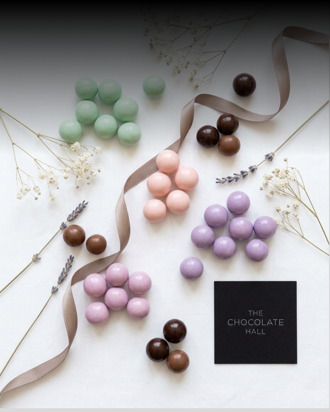 Colorful chocolate spheres in pastel pink, purple, green, brown, and beige arranged with dried flowers and a black sign that reads 'The Chocolate Hall' on a white background.