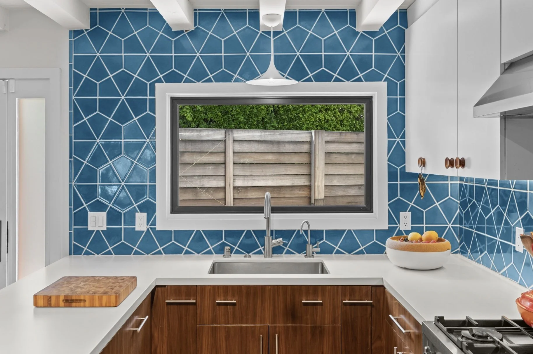 Modern kitchen with geometric blue tile backsplash, white cabinets, wooden lower cabinets, stainless steel sink, and a window overlooking a wooden fence and greenery.