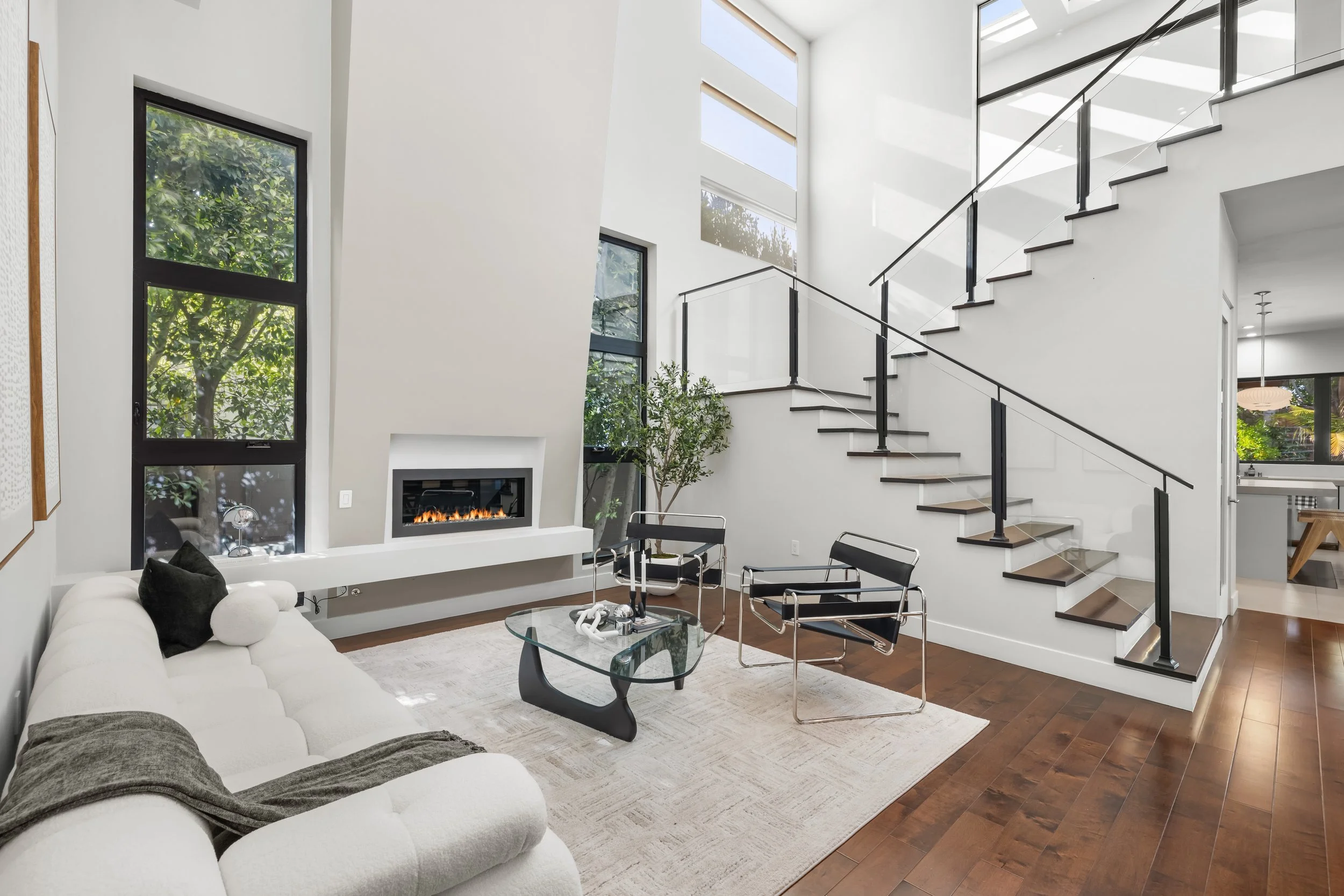 Modern living room with white walls and a high ceiling, featuring a white sofa, two black and chrome chairs, a glass coffee table, a fireplace, large windows, a staircase with black railings, and hardwood floors.