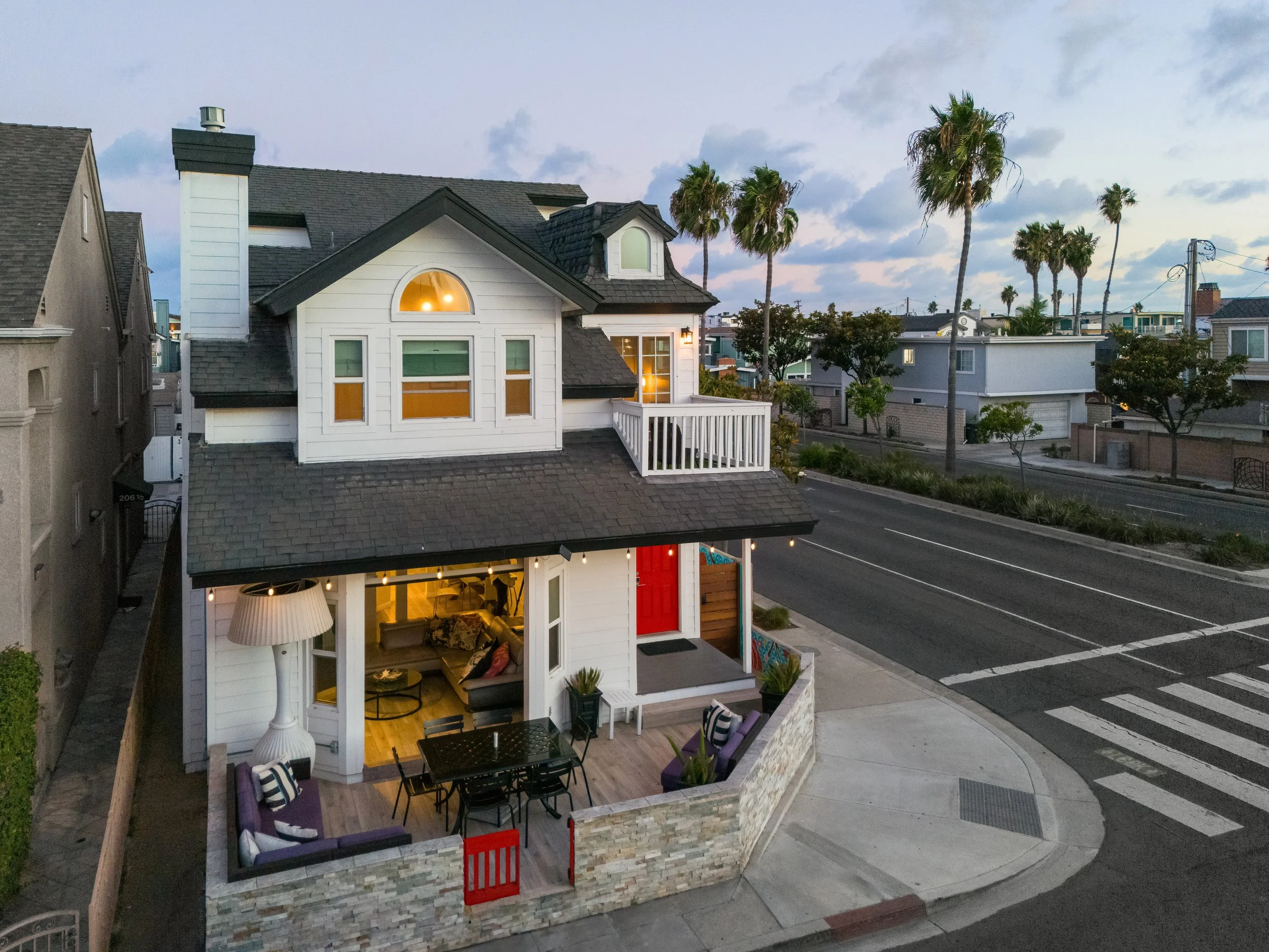 A two-story white house with a small balcony, large windows with colorful blinds, and warm interior lighting. The house features a gray shingled roof and multiple palm trees in the background, suggesting a coastal or Southern California neighborhood.