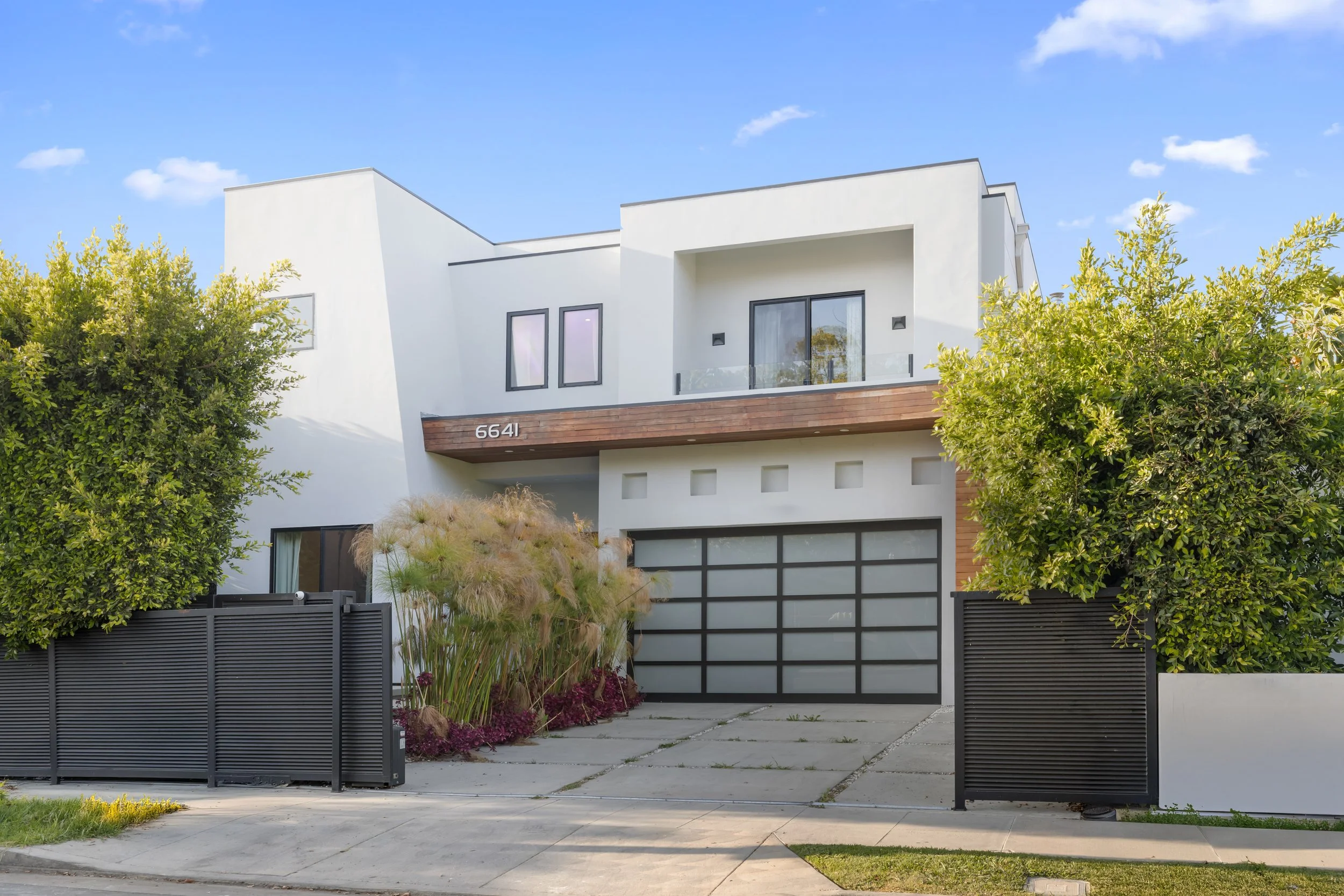 Modern white house with large windows and a garage, surrounded by greenery and trees, under a blue sky.