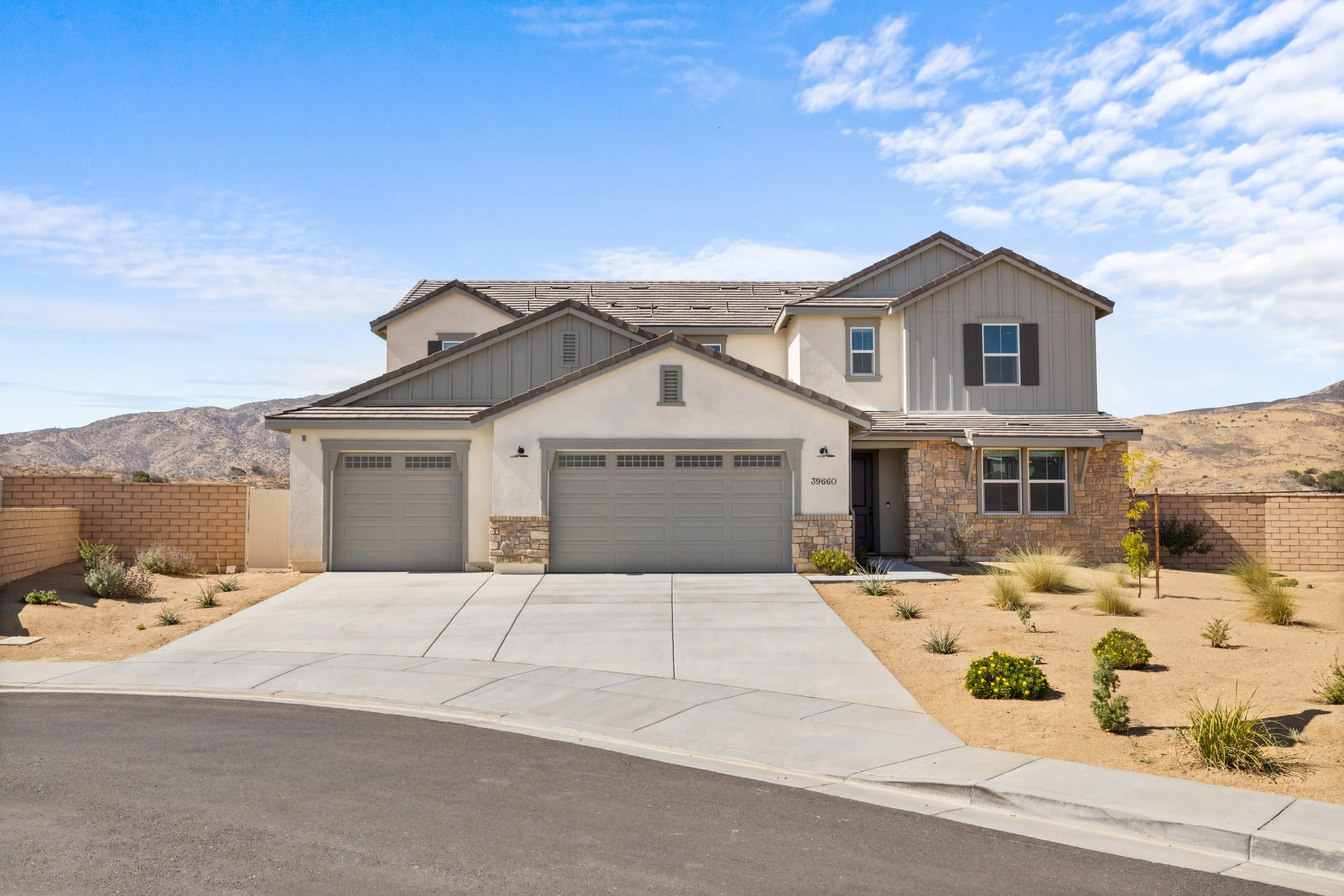 A two-story suburban house with a three-car garage, beige and gray exterior, and a desert landscaping yard.
