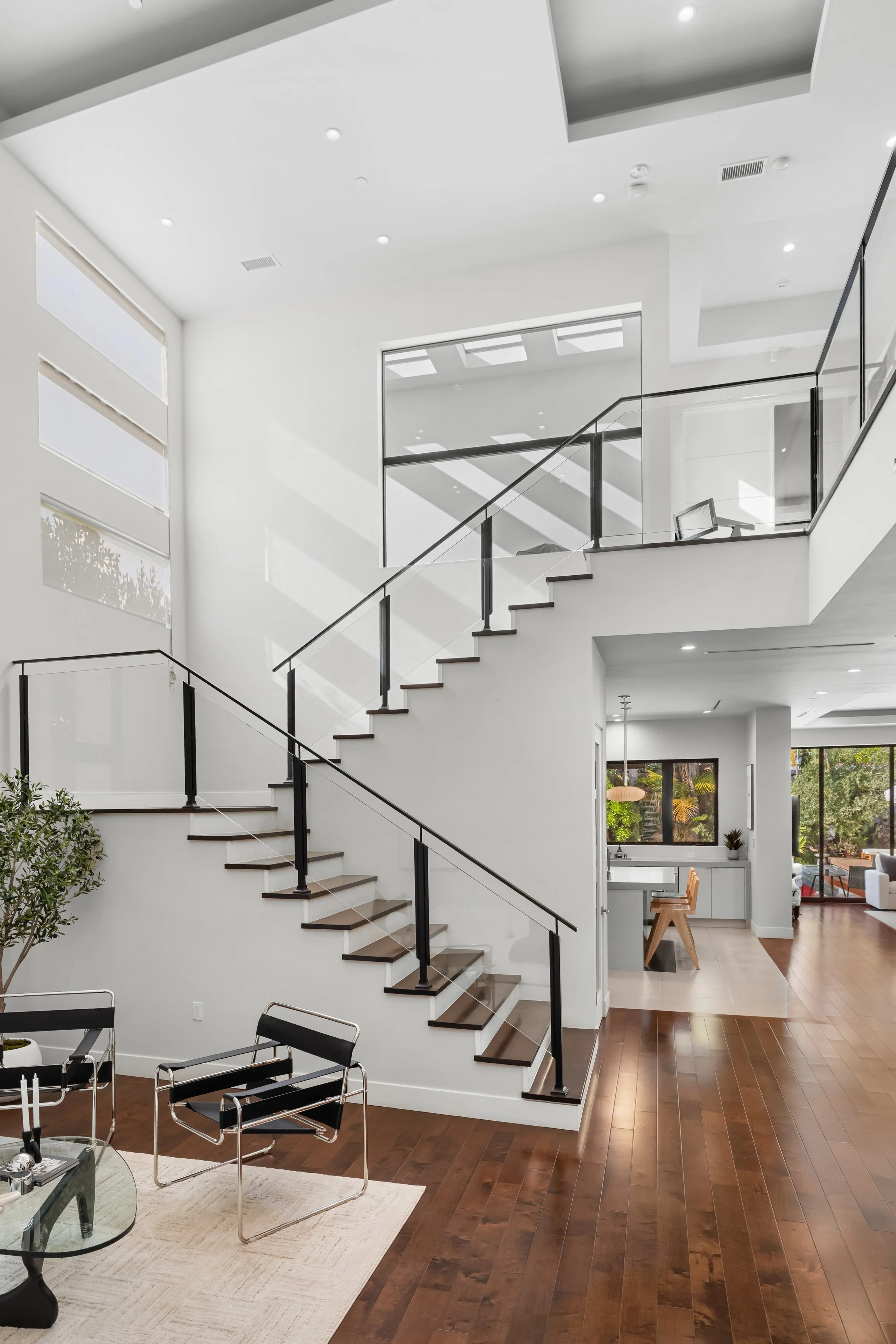 Modern interior home with white walls, wooden flooring, staircase with black railing, and large windows letting in natural light.
