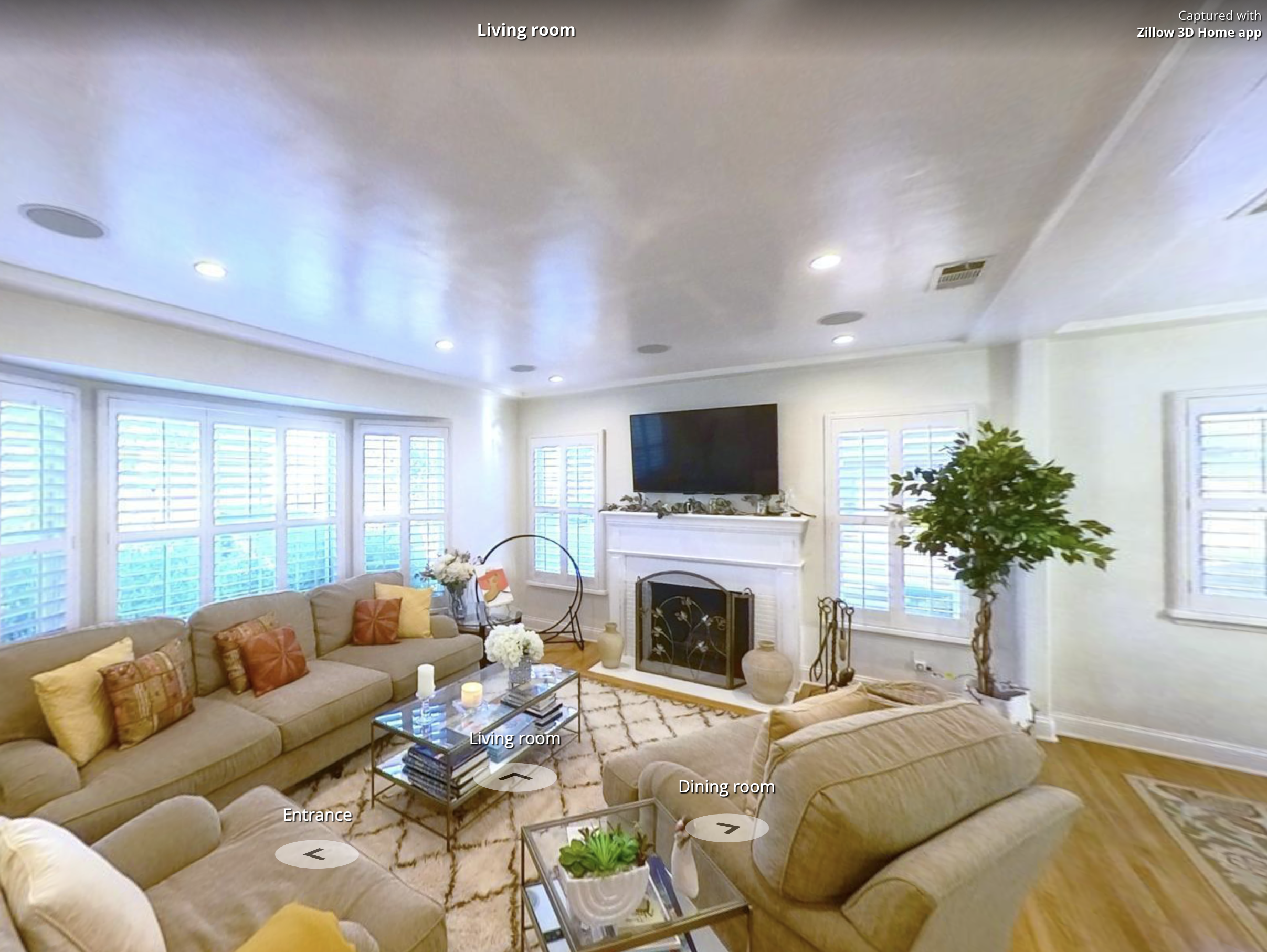 Bright living room with beige sectional sofas, colorful throw pillows, a glass coffee table with candles and flowers, multiple large windows with shutters, a fireplace with decorations, a large flat-screen TV above the fireplace, a potted plant, and