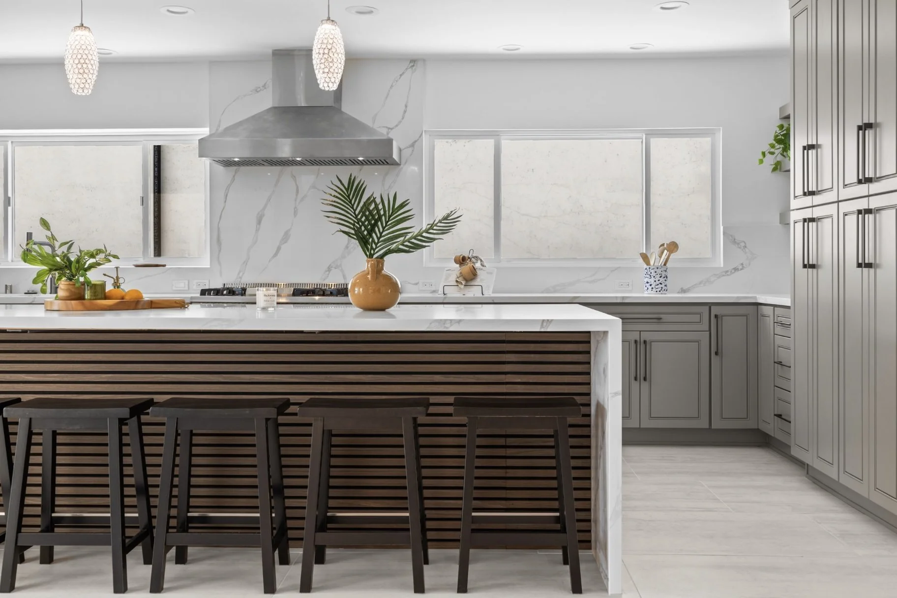 Modern kitchen with marble countertops, light gray cabinetry, black stools, and decorative plants.