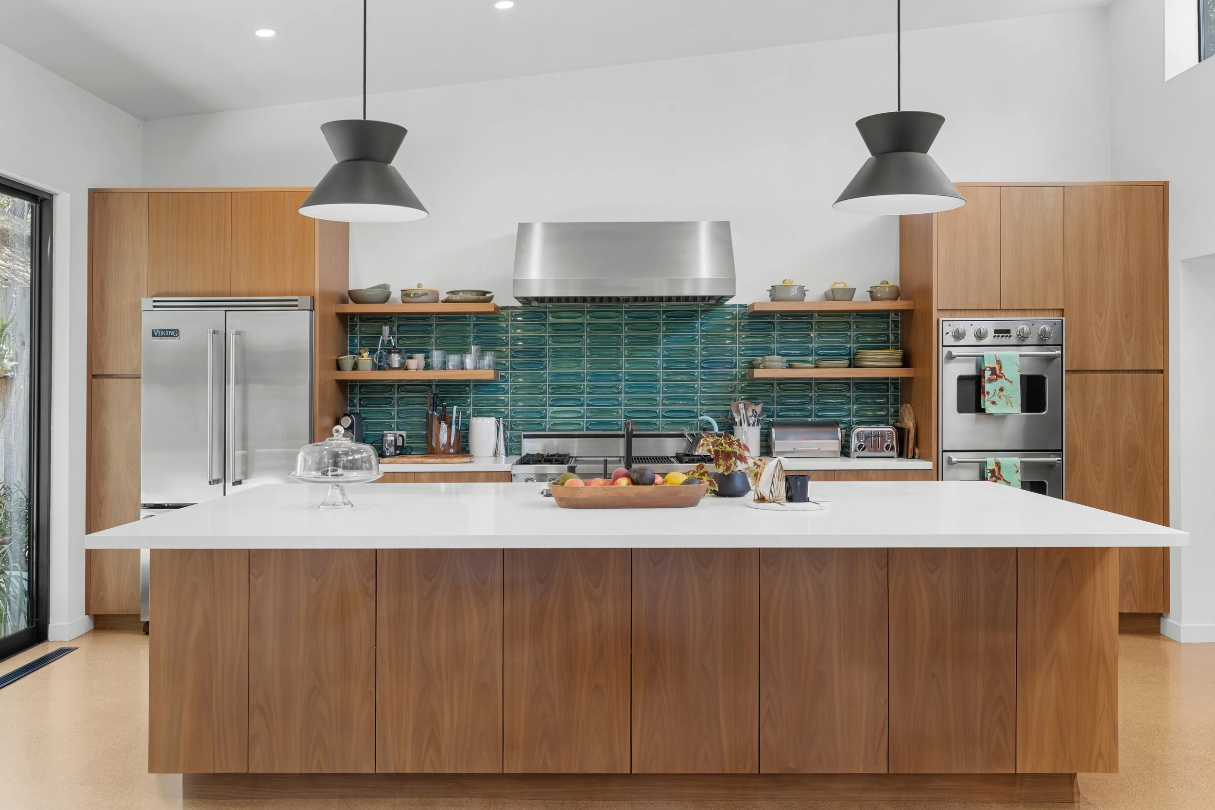 Modern kitchen with wooden cabinets, a white island, teal tile backsplash, stainless steel appliances, open shelves with dishes, and black pendant lights.