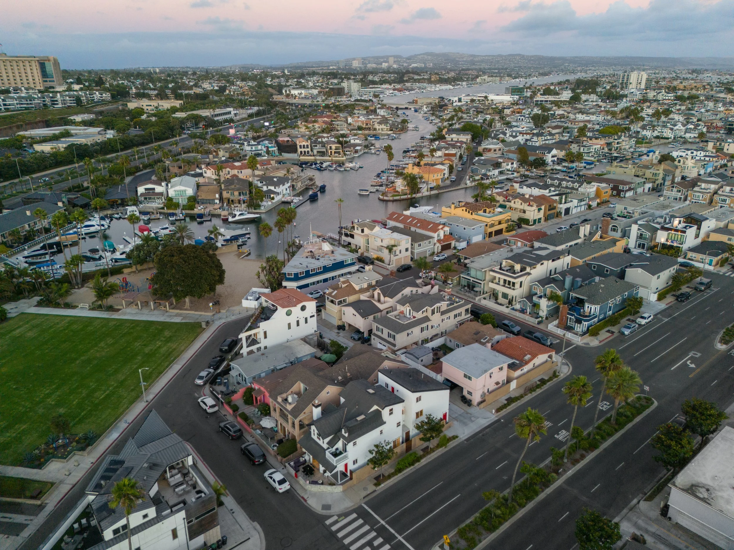 Aerial view of a coastal residential neighborhood with houses, a canal filled with boats, and a park with a playground, palm trees, and a large grassy area.