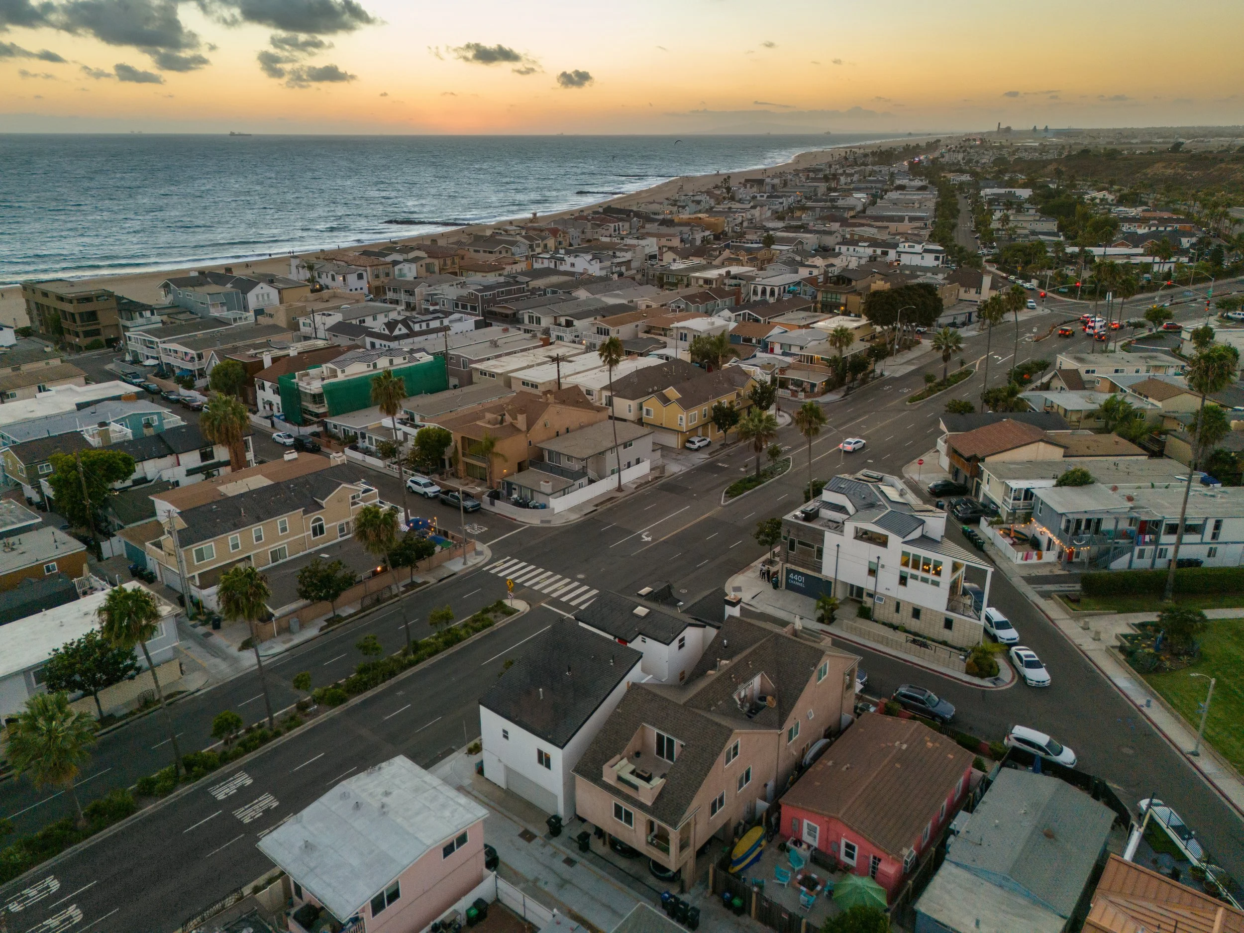 Aerial view of a beachside neighborhood at sunset with ocean, sandy beach, and houses with palm trees.
