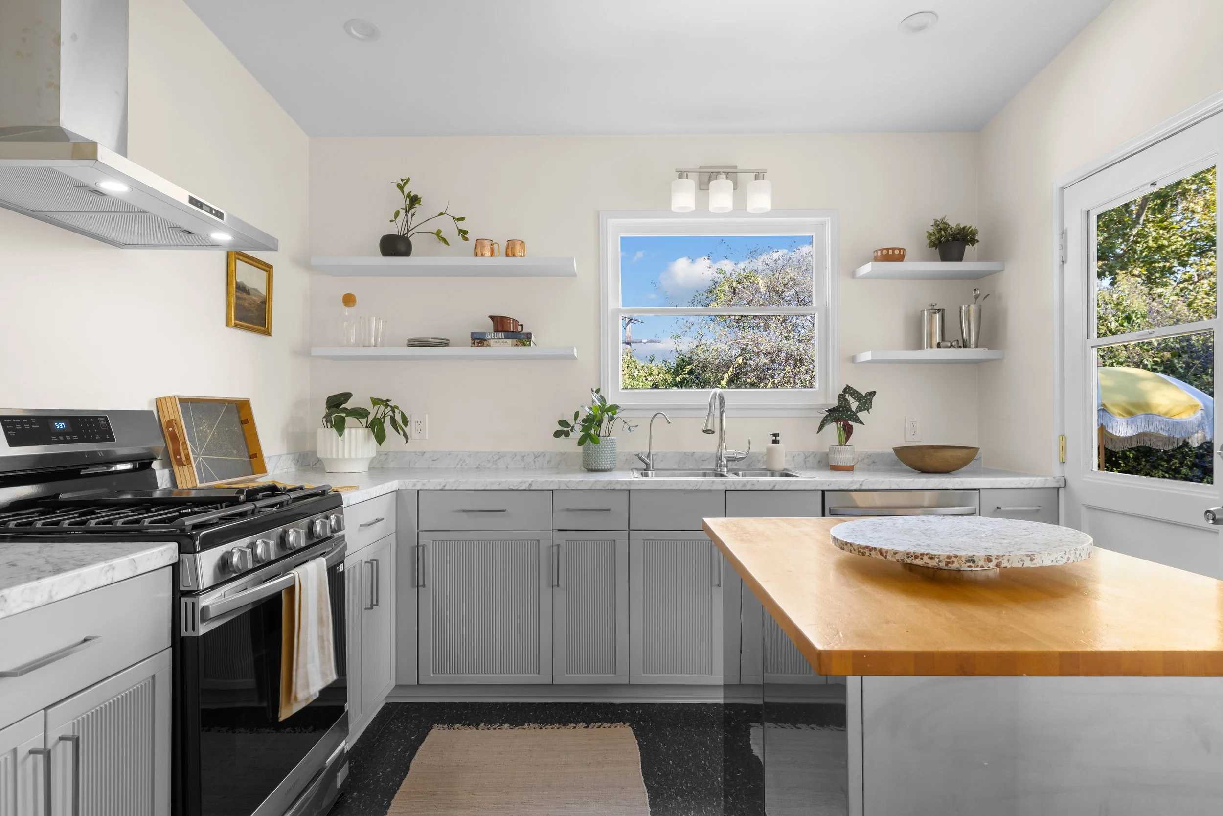 Bright kitchen with white walls, gray cabinets, marble countertops, and open shelves. Includes a stove, plants, and a large window showing a blue sky and trees.