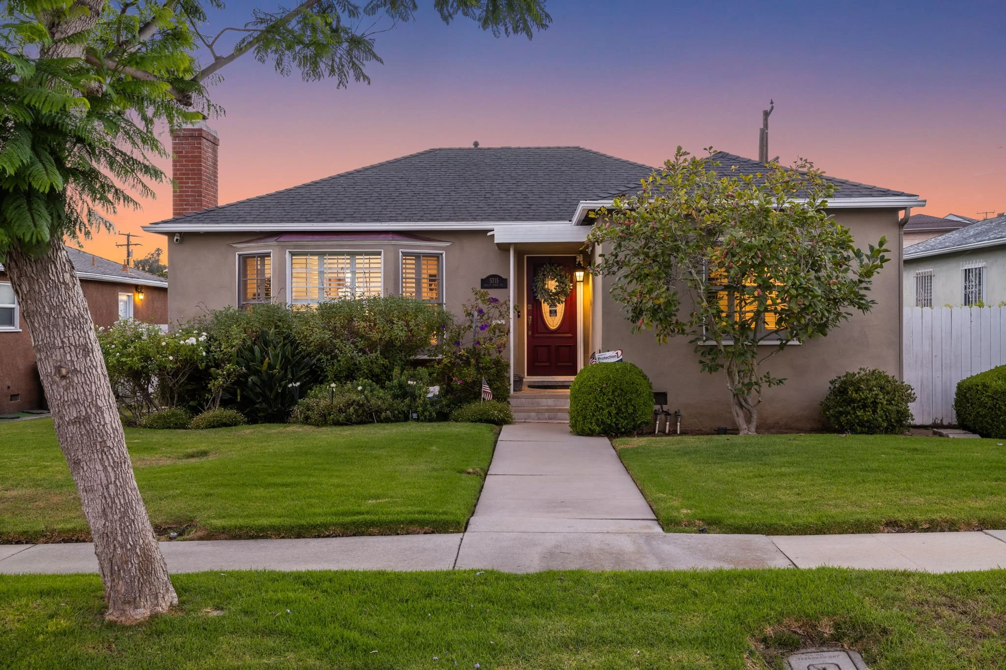Front view of a single-story house with a well-manicured lawn, trees, and bushes during sunset. The house has a central door with a decorative wreath and illuminated windows.
