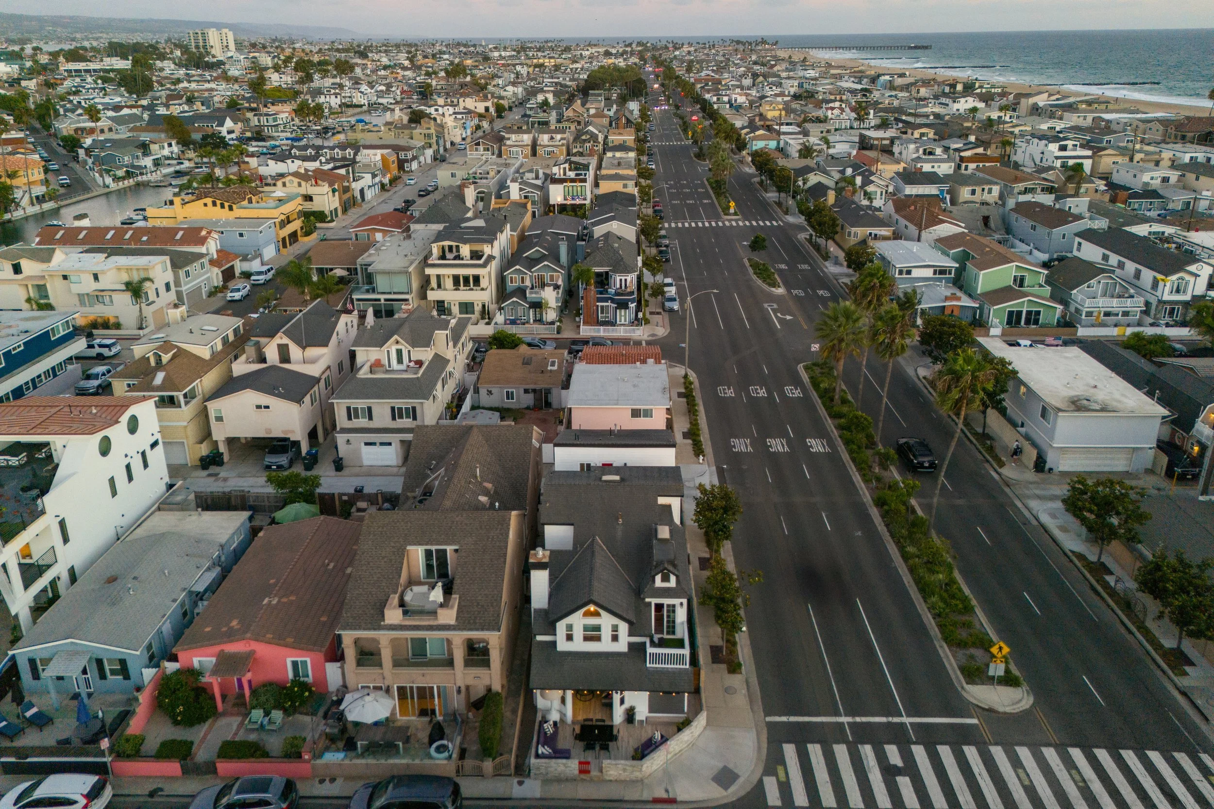 Aerial view of a coastal neighborhood with closely packed houses, a multi-lane street, palm trees, and the ocean in the background.