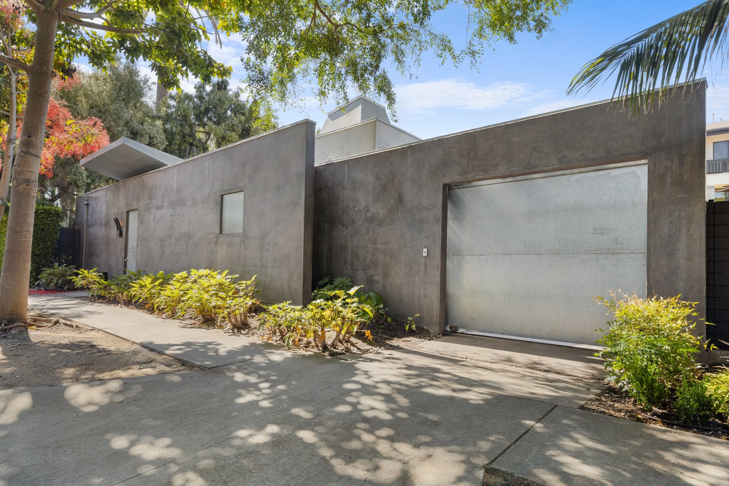 Modern gray concrete house with a garage door, small windows, surrounded by trees and shrubs on a sunny day.