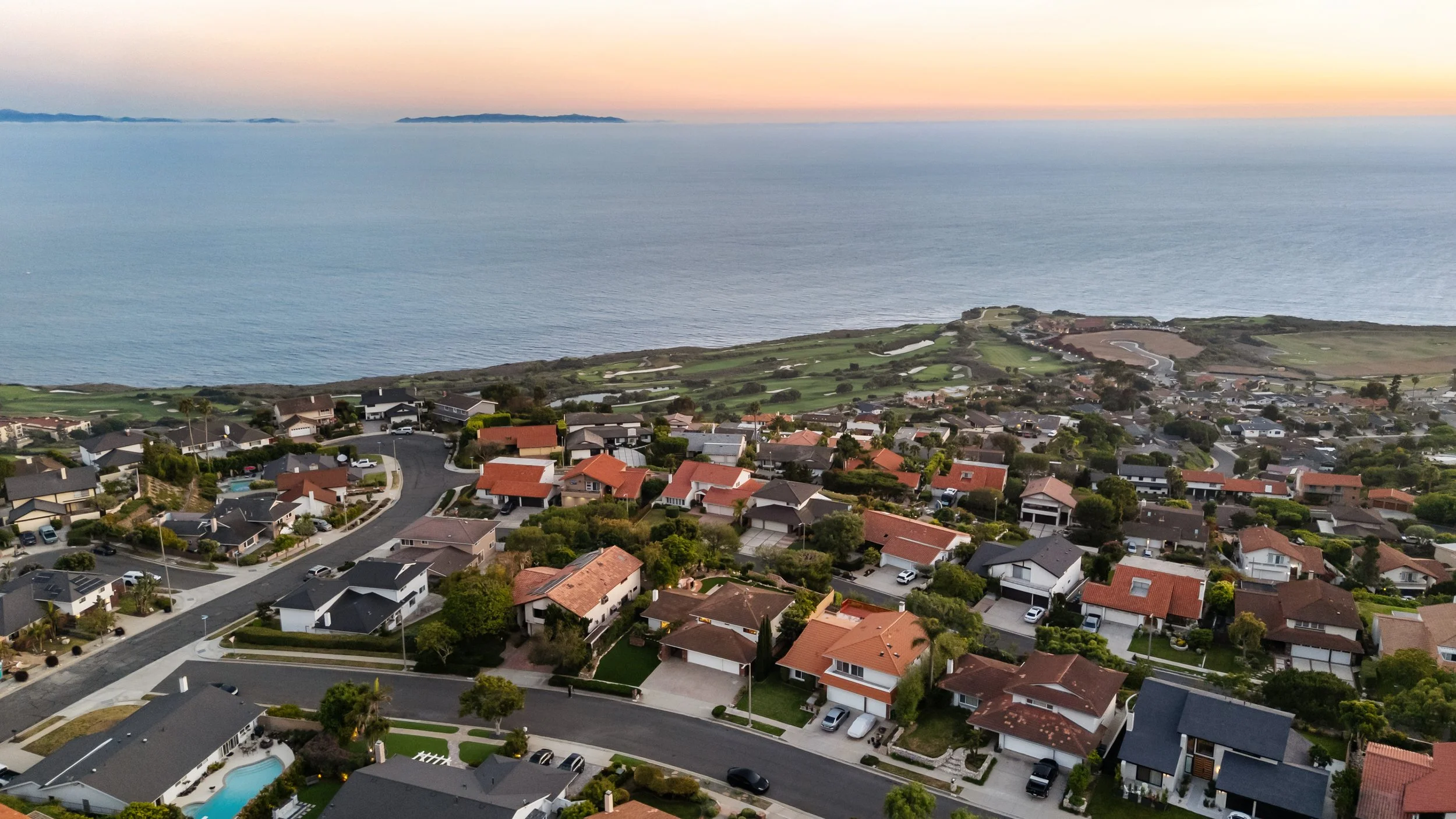 Aerial view of a coastal suburban neighborhood with houses, streets, trees, and a nearby body of water during sunset.